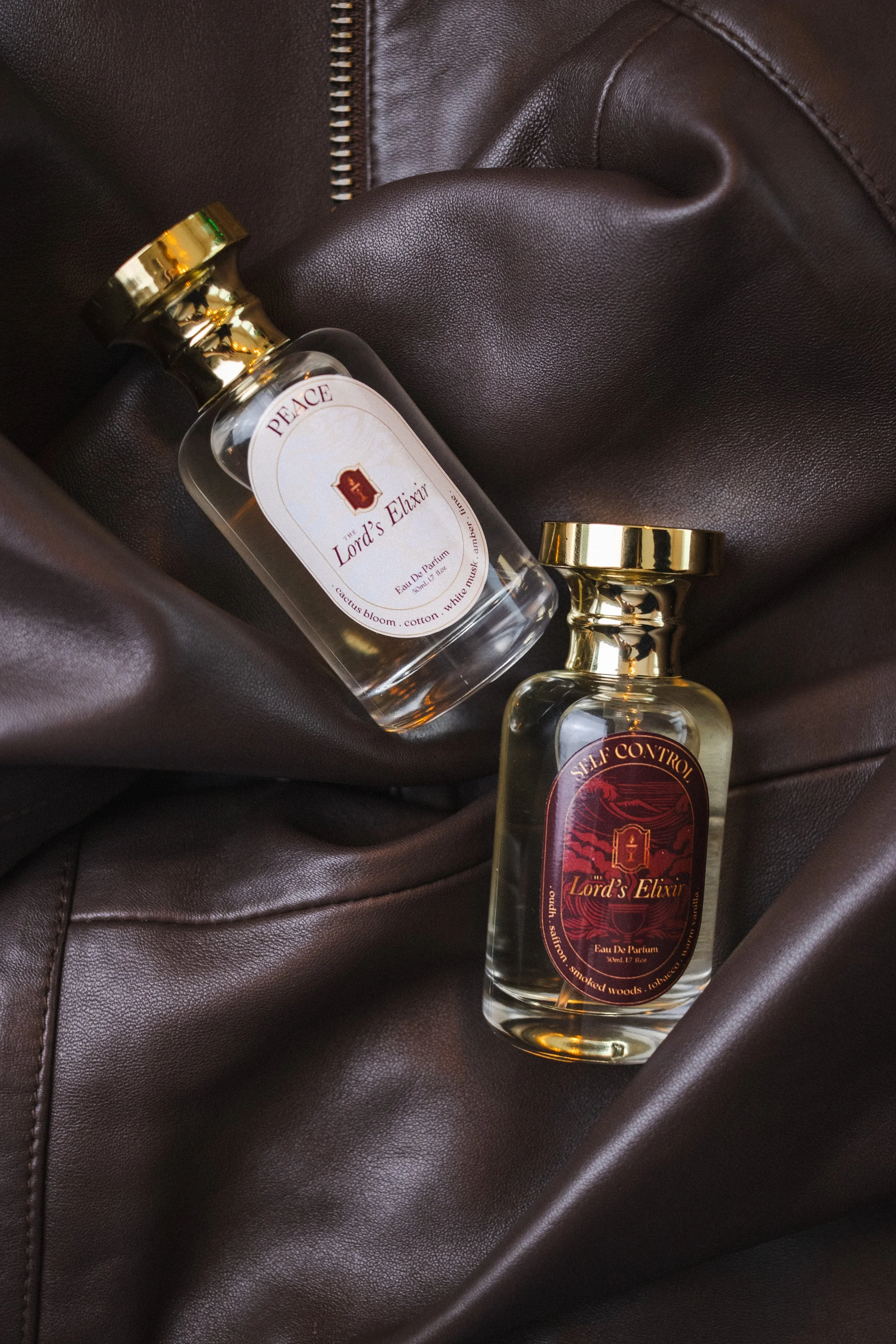 Two bottles of perfume, one lying on a dark brown leather surface, and the other resting upright, both labeled 'Lord's Elixir' with gold caps and decorative labels.