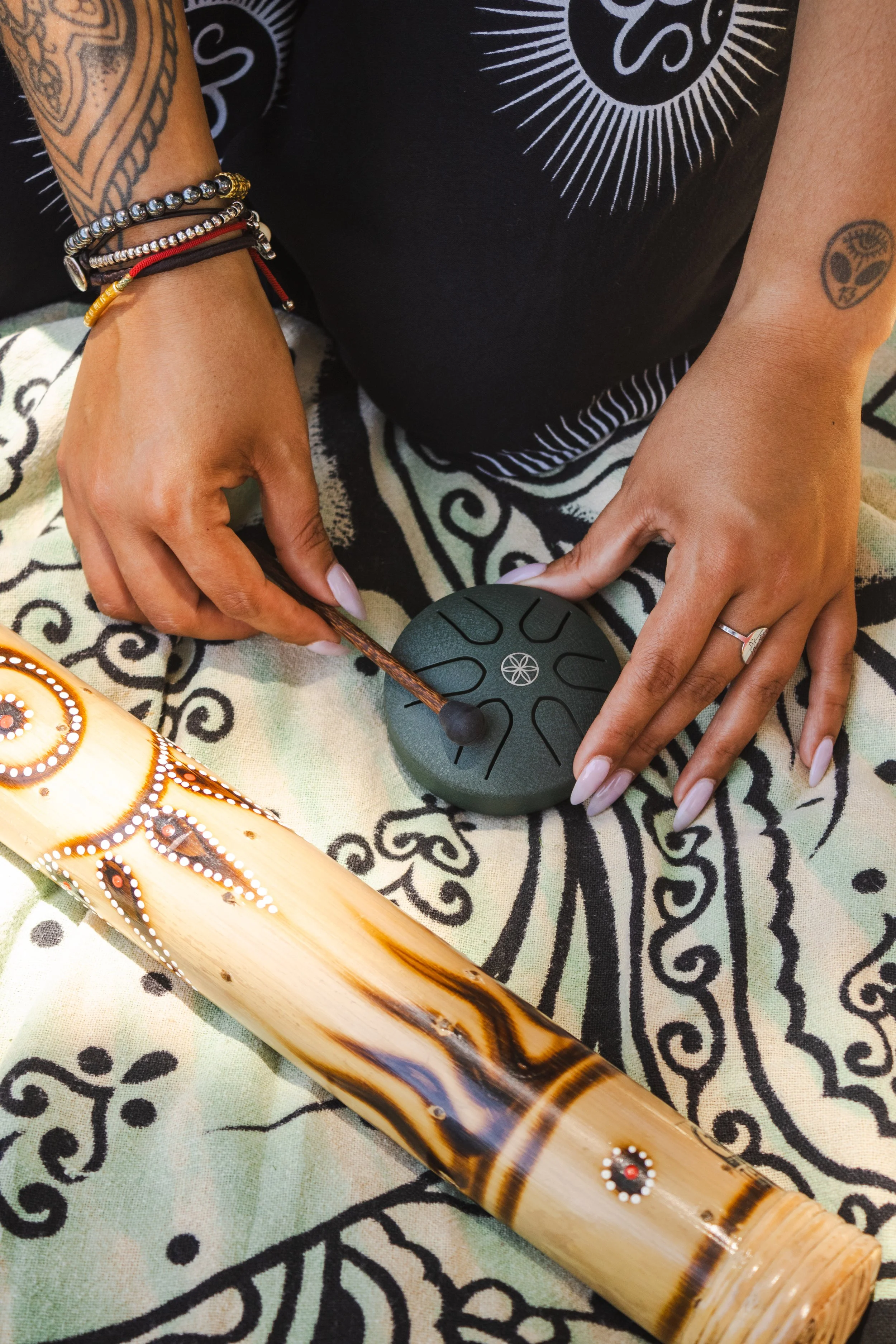 A person playing a small instrument with a mallet, surrounded by tribal jewelry and tattoos, on a cloth with tribal patterns, with a decorated wooden instrument nearby.