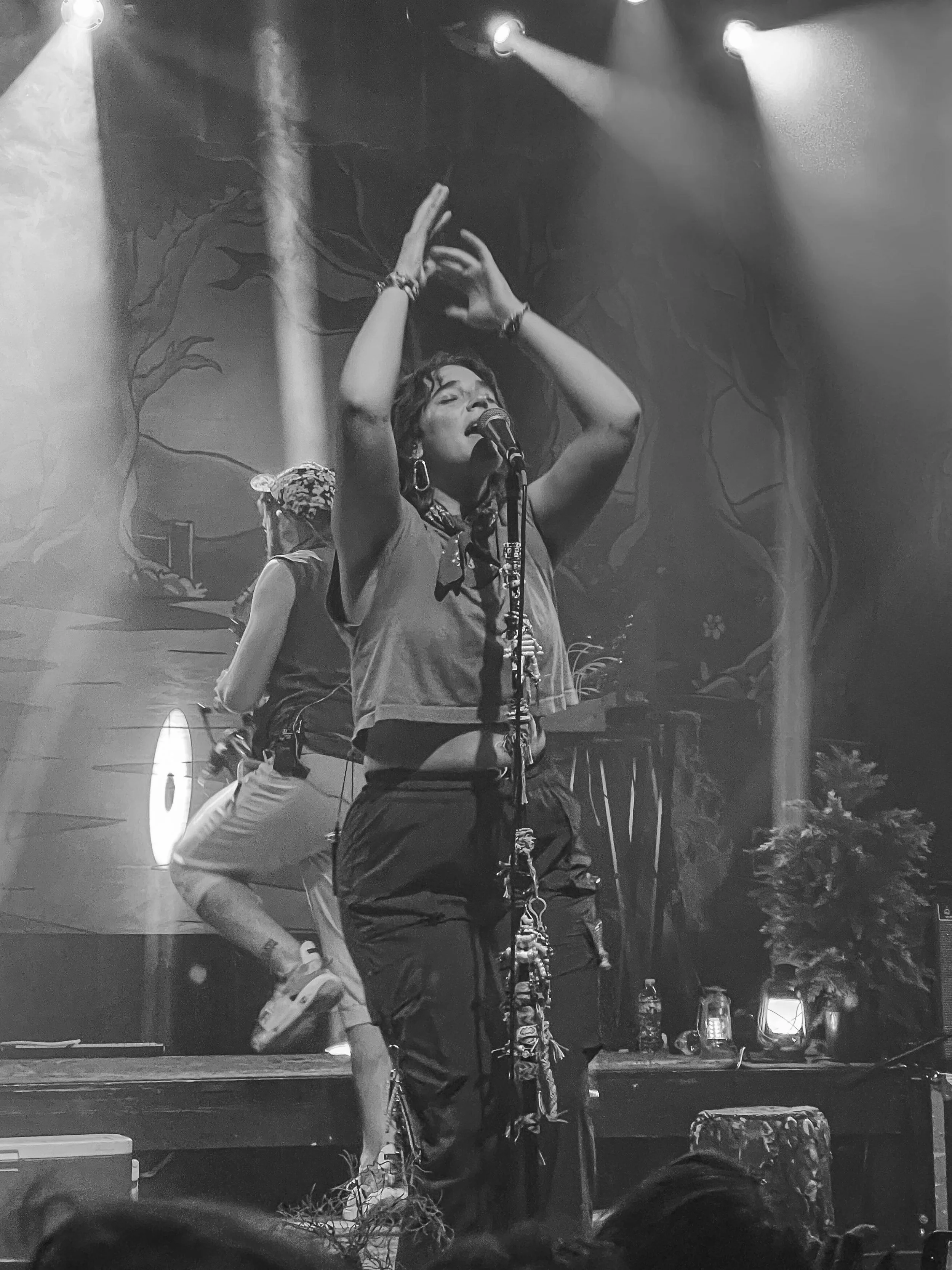 A woman singing on stage with a microphone, making expressive gestures with her hands, during a performance with stage lights and backdrop behind her.