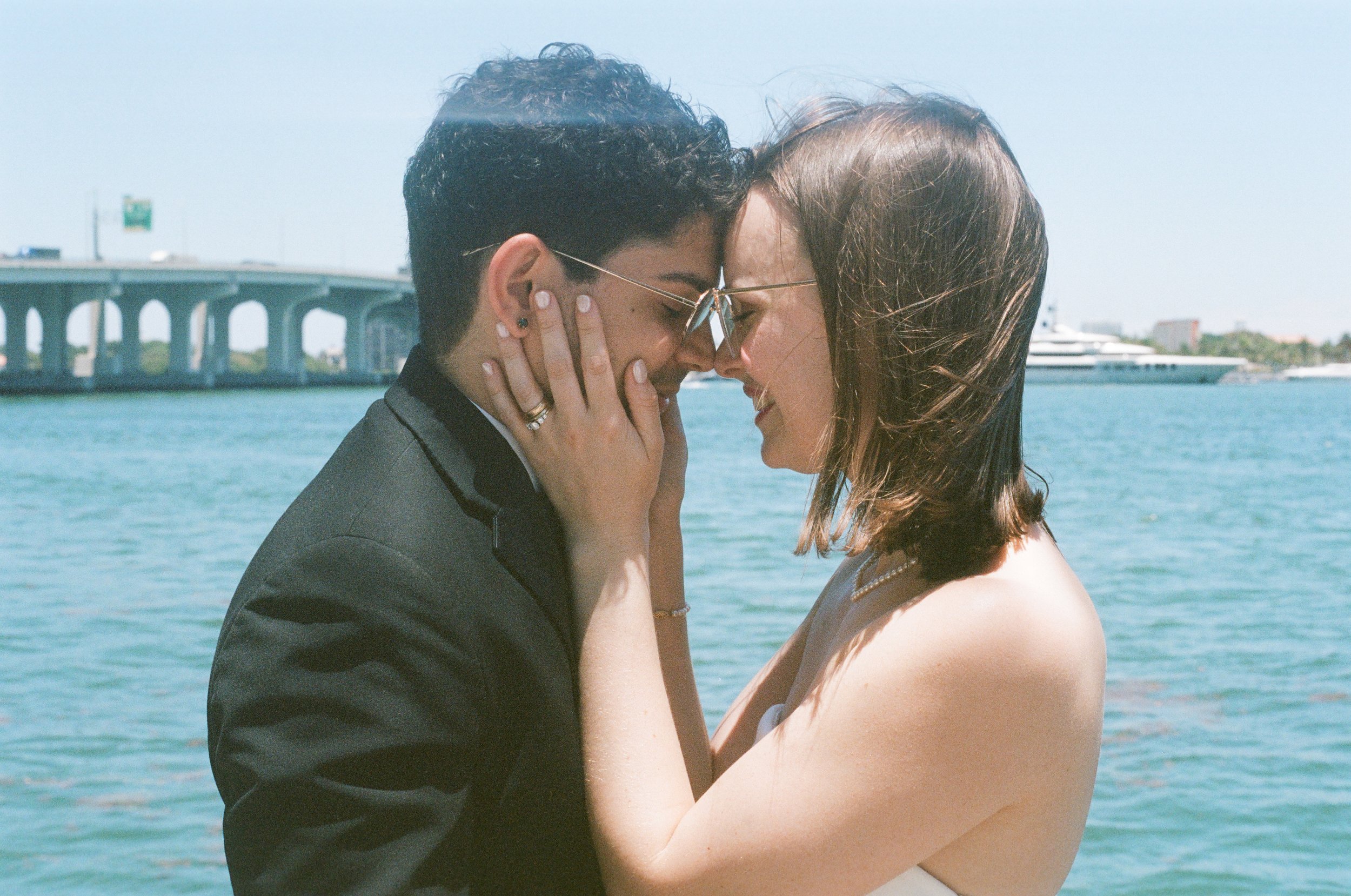 A couple is embracing and touching foreheads near a body of water with a bridge and boats in the background.