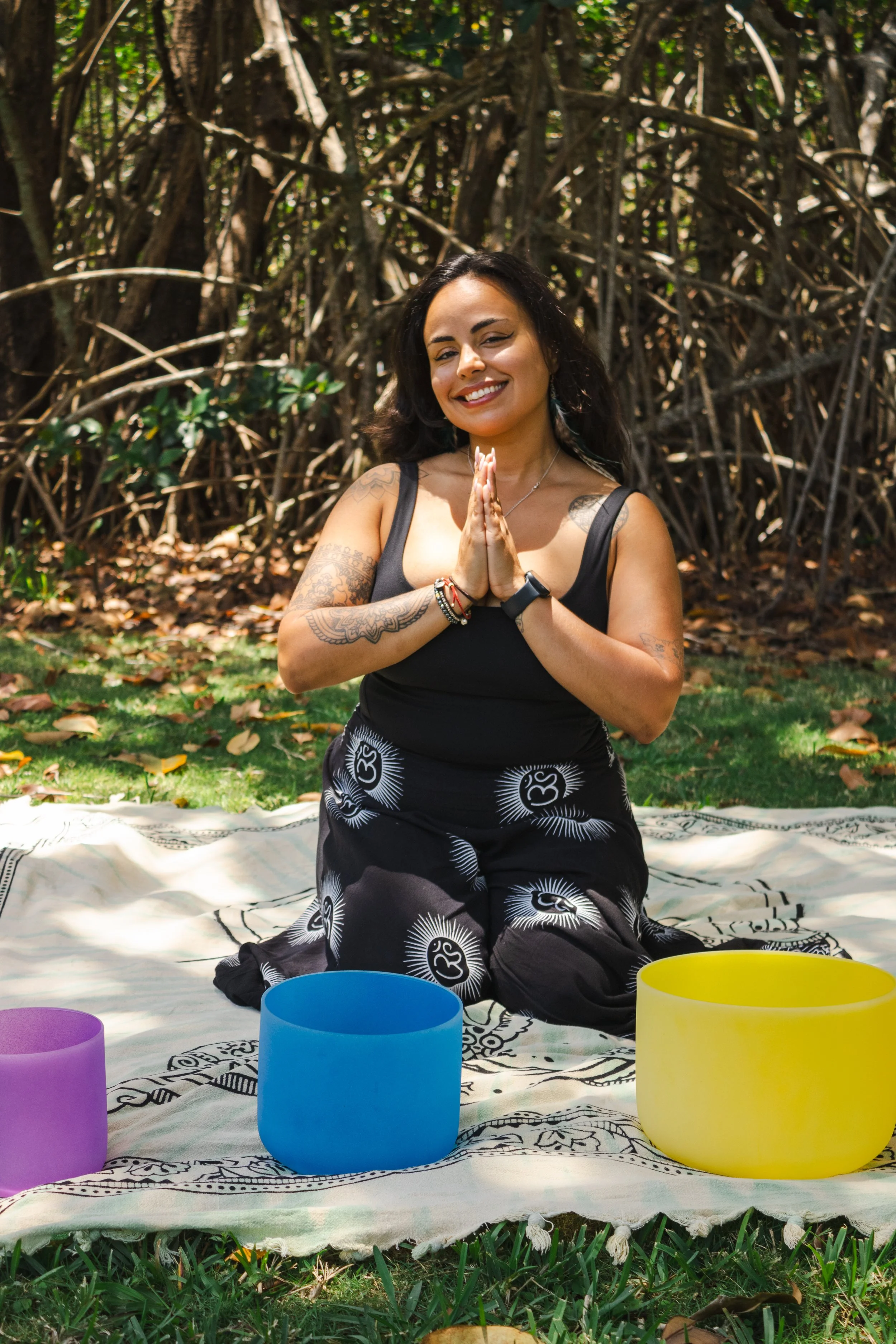A woman practicing meditation outdoors on a sunny day, sitting cross-legged on a patterned cloth on the grass, with her hands in prayer position and smiling. Colorful singing bowls are placed in front of her, and a background of trees and foliage sur