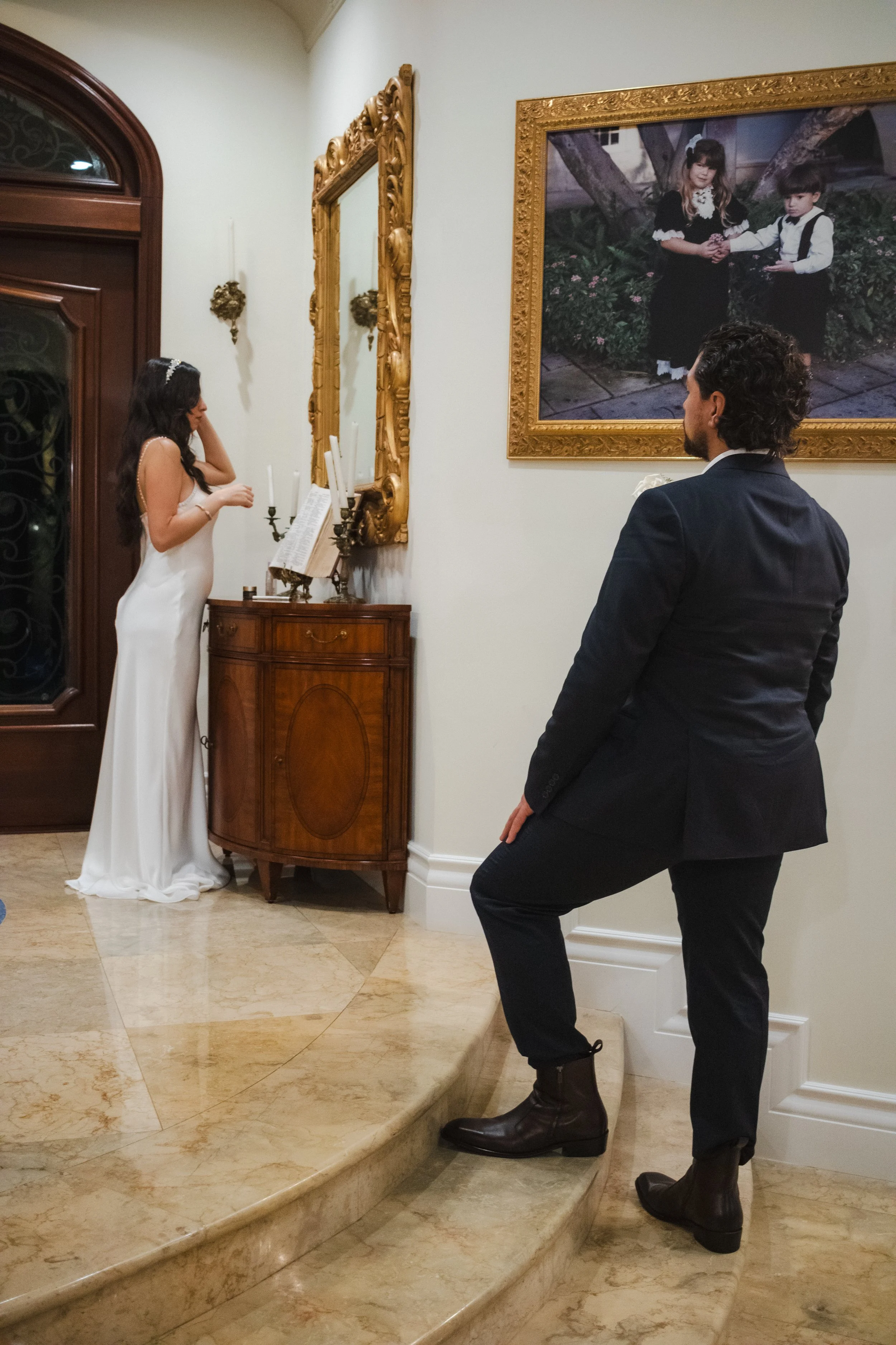 A woman in a white dress is standing at a small table reading from a book, with a man in dark clothing and boots looking at her. There is a large framed photo of two children holding hands on the wall.