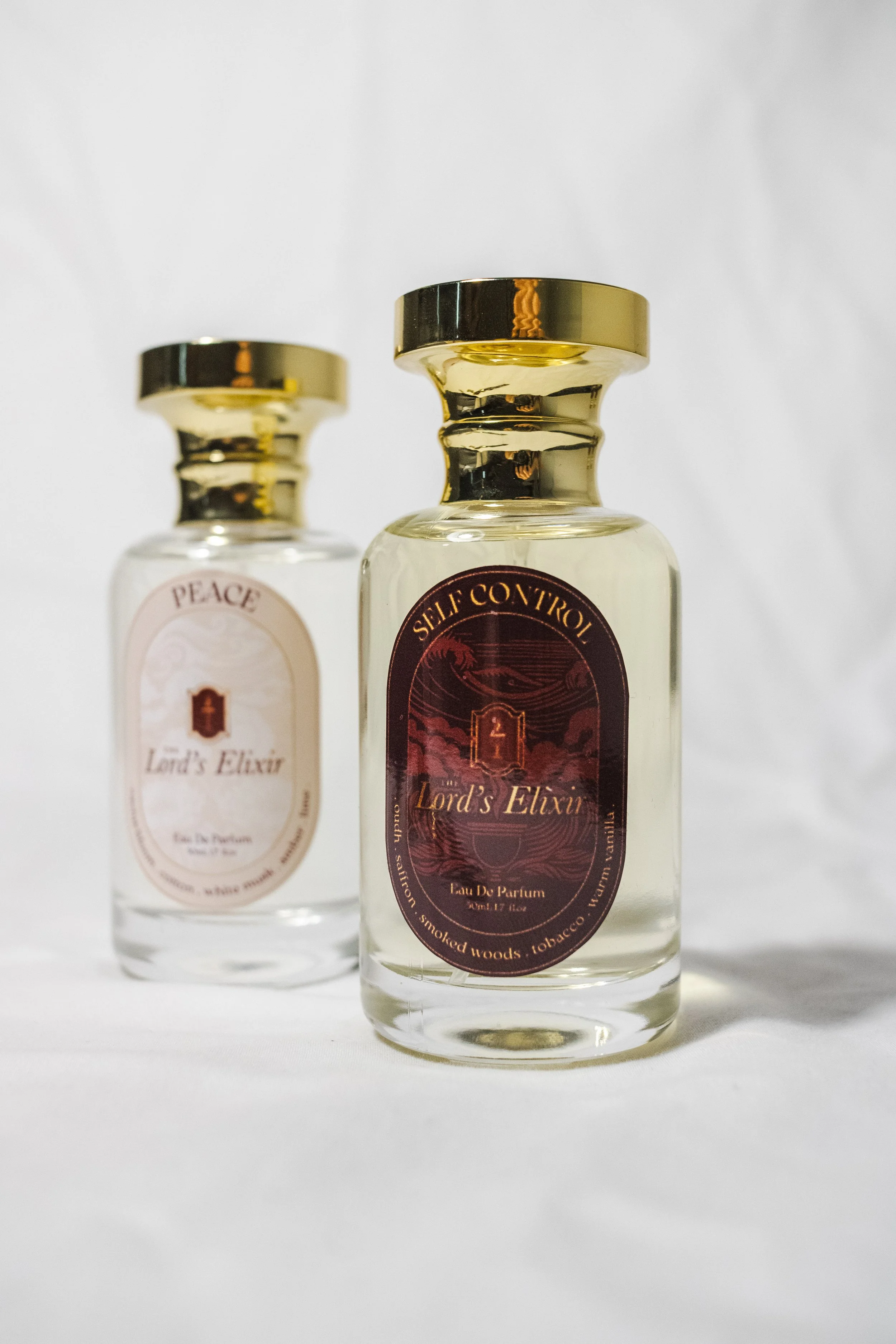 Two small perfume bottles with gold caps, labeled 'Peace' and 'Self Control,' standing on a white surface against a plain background.