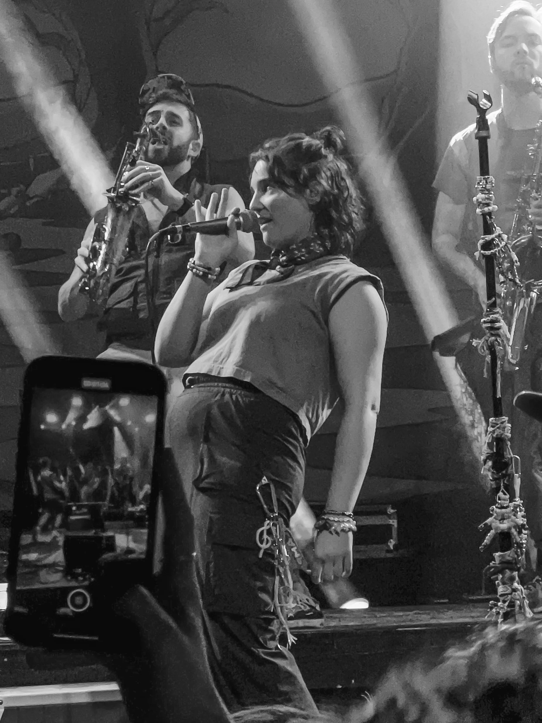 Black and white photo of a woman singing into a microphone with people and instruments around her on a stage.