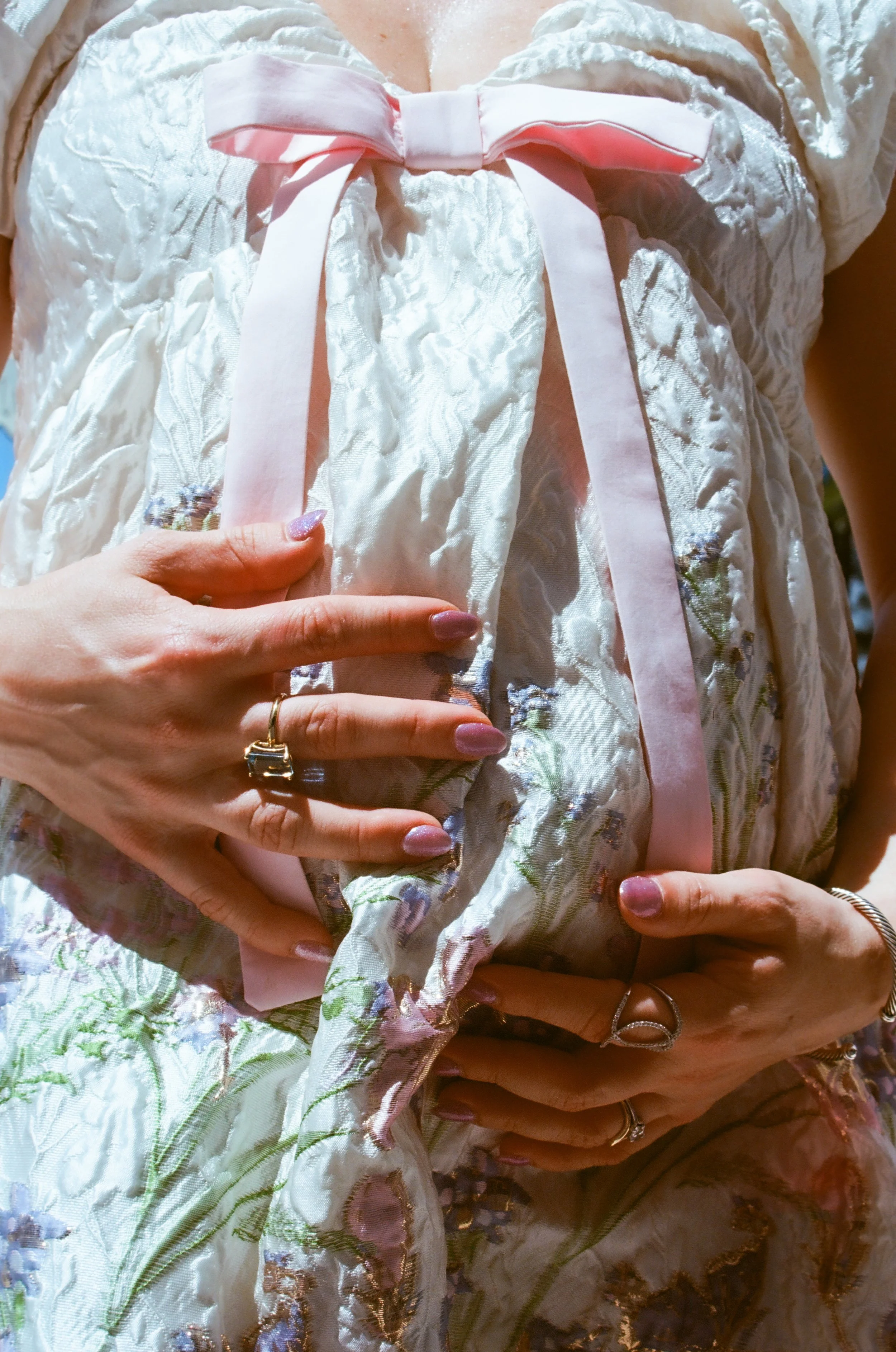 Close-up of a person wearing an elegant, embroidered white dress with floral patterns and a pink satin ribbon tied in a bow at the chest. The person is holding their hands over their stomach, displaying rings on their fingers and painted nails.
