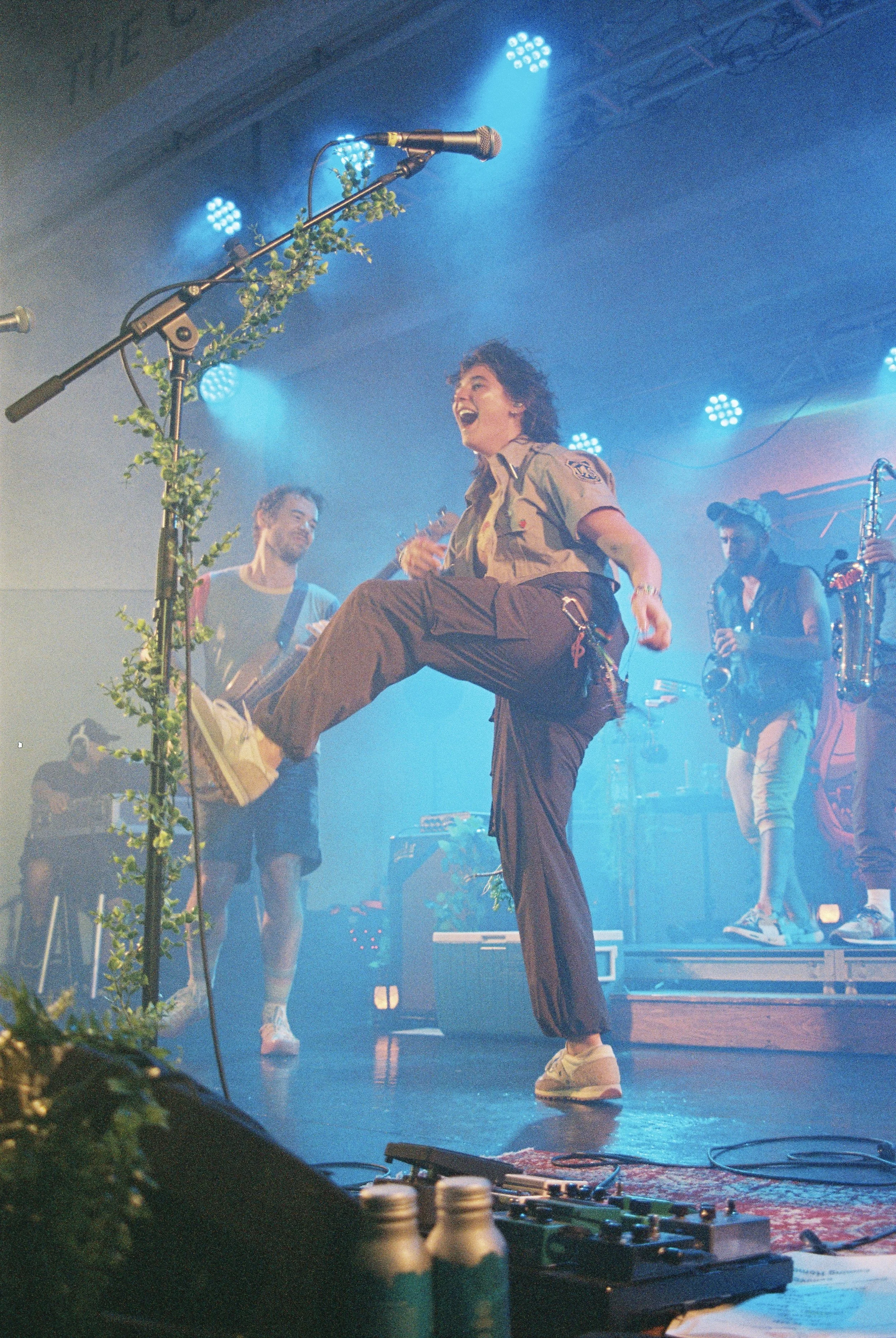 A woman in a military-style shirt and cargo pants performing on stage, raising one leg while holding her arm in the air, with a microphone stand decorated with green leaves nearby. Two musicians are seen in the background, one playing a guitar and th