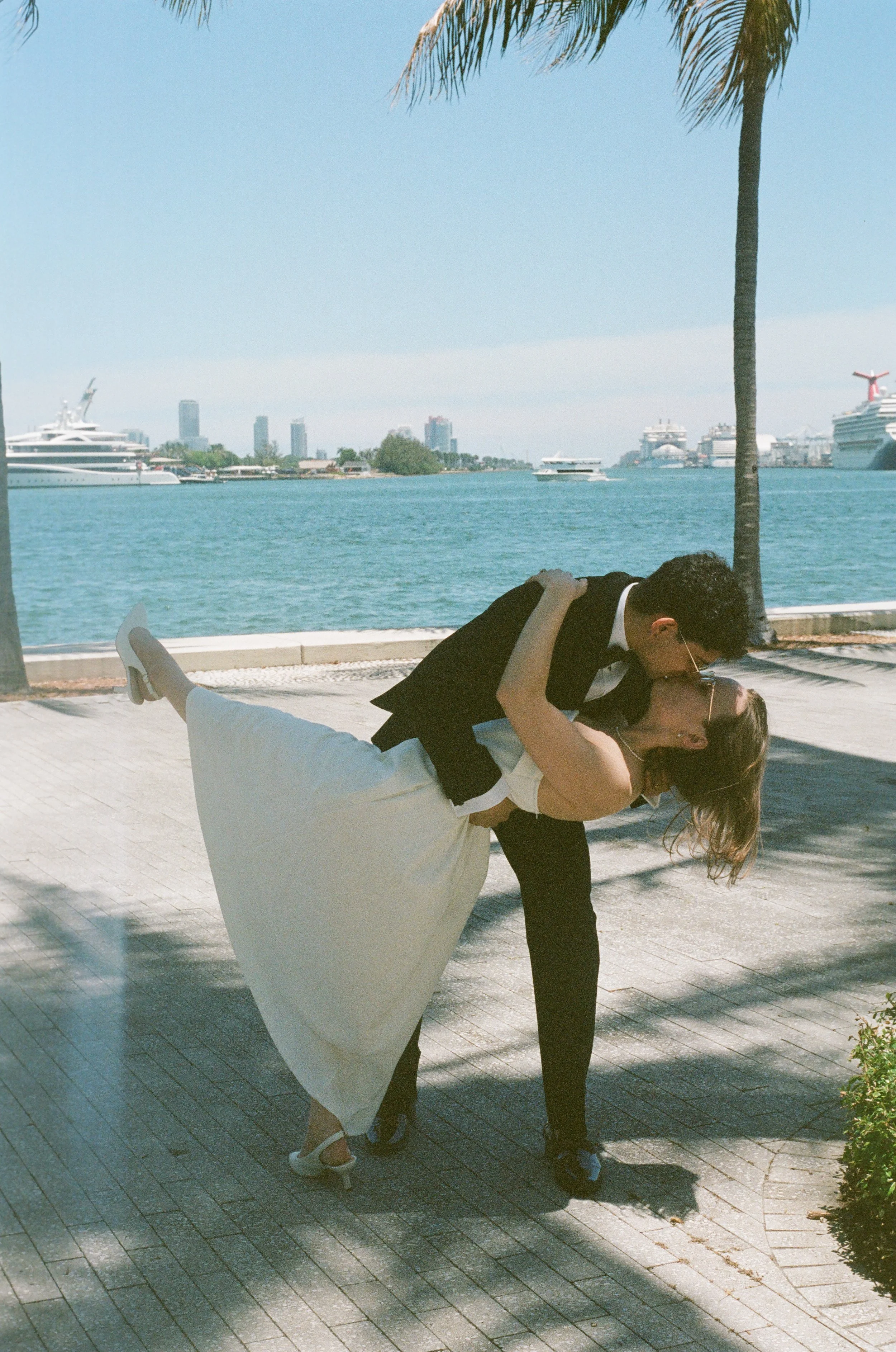 A couple dressed in formal wedding attire sharing a dance outdoors near a waterfront with cruise ships and a city skyline in the background.