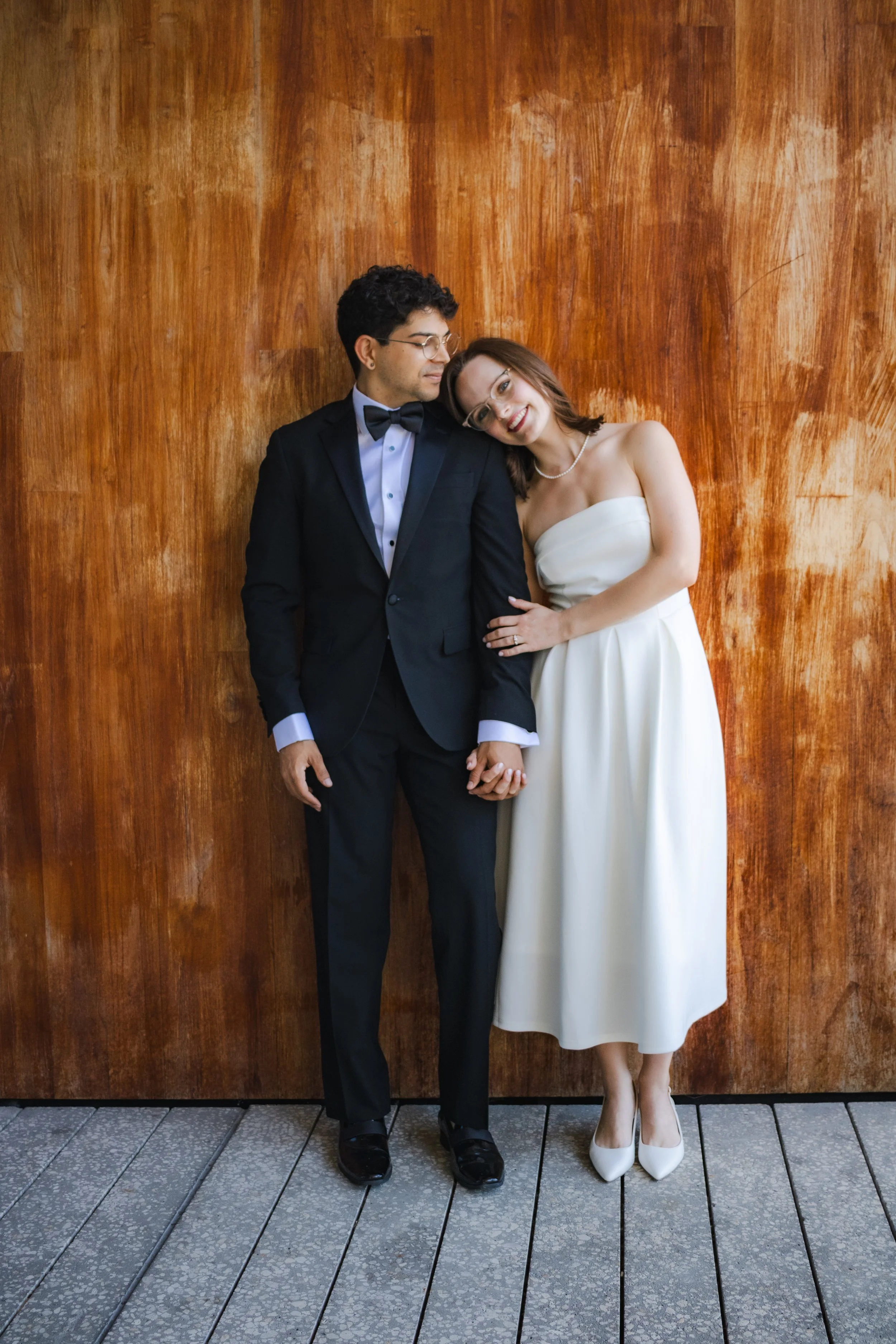 A man and woman dressed in formal wedding attire standing close together, holding hands, against a wooden wall background. The man is wearing a black tuxedo with a bow tie, and the woman is wearing a strapless white wedding dress and high heels. The 