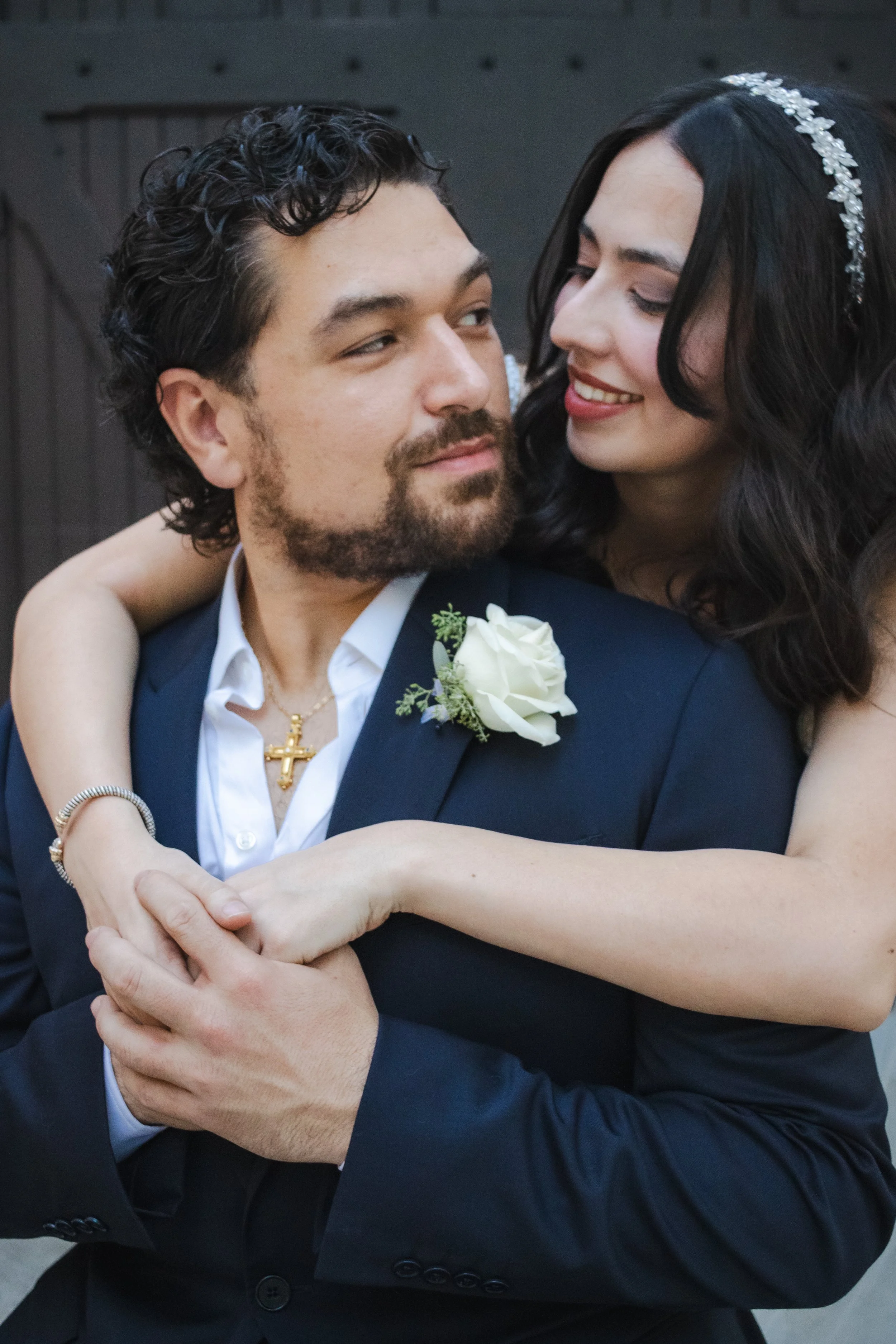 A couple dressed in wedding attire, the man wearing a navy blue suit with a white shirt and gold chain necklace with a cross, and the woman wearing a headband with floral details, smiling and holding hands.