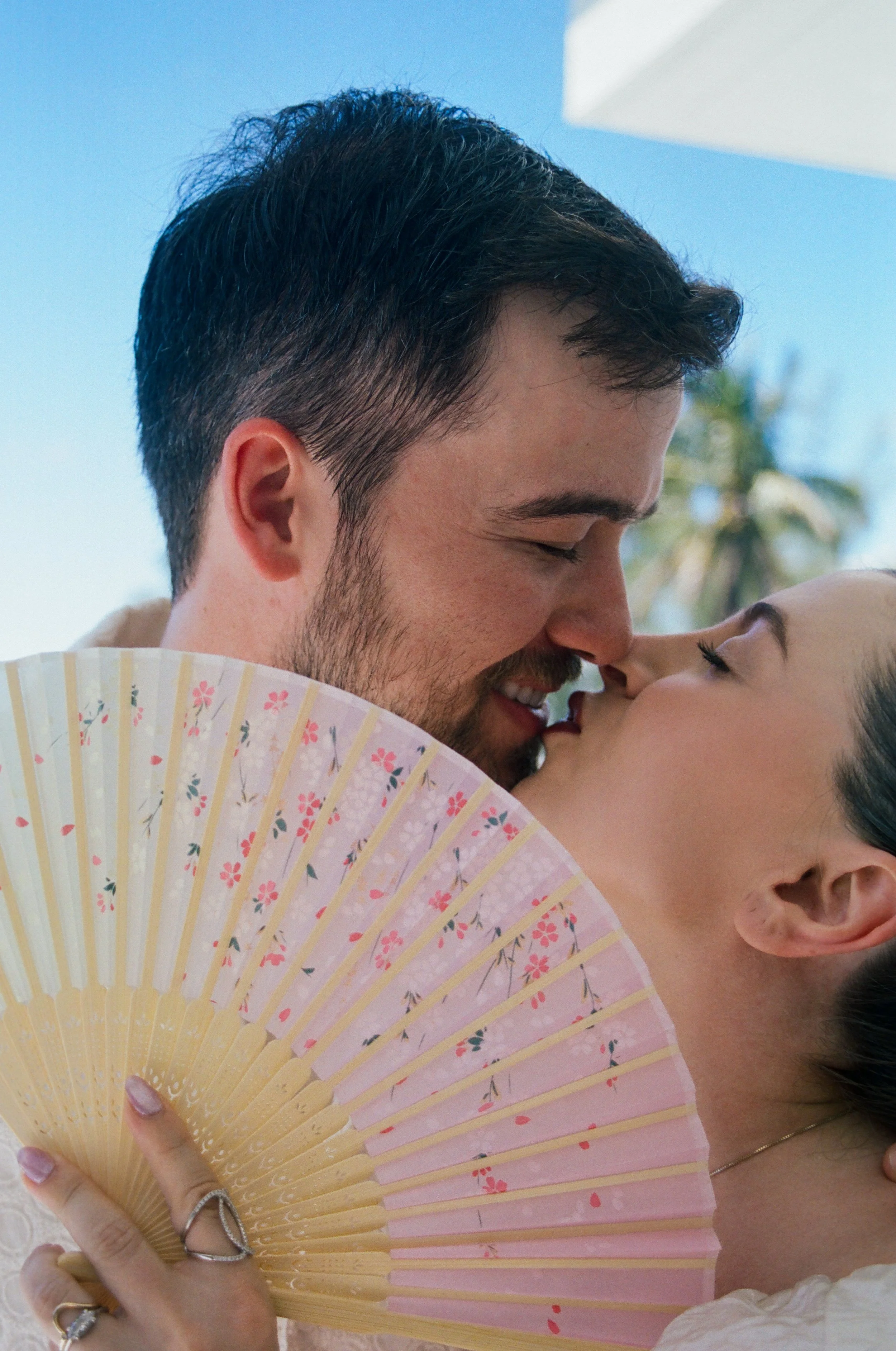 A close-up of a man and woman sharing an affectionate moment, with the man smiling and the woman gently touching his face, partially obscured by a pink floral fan.