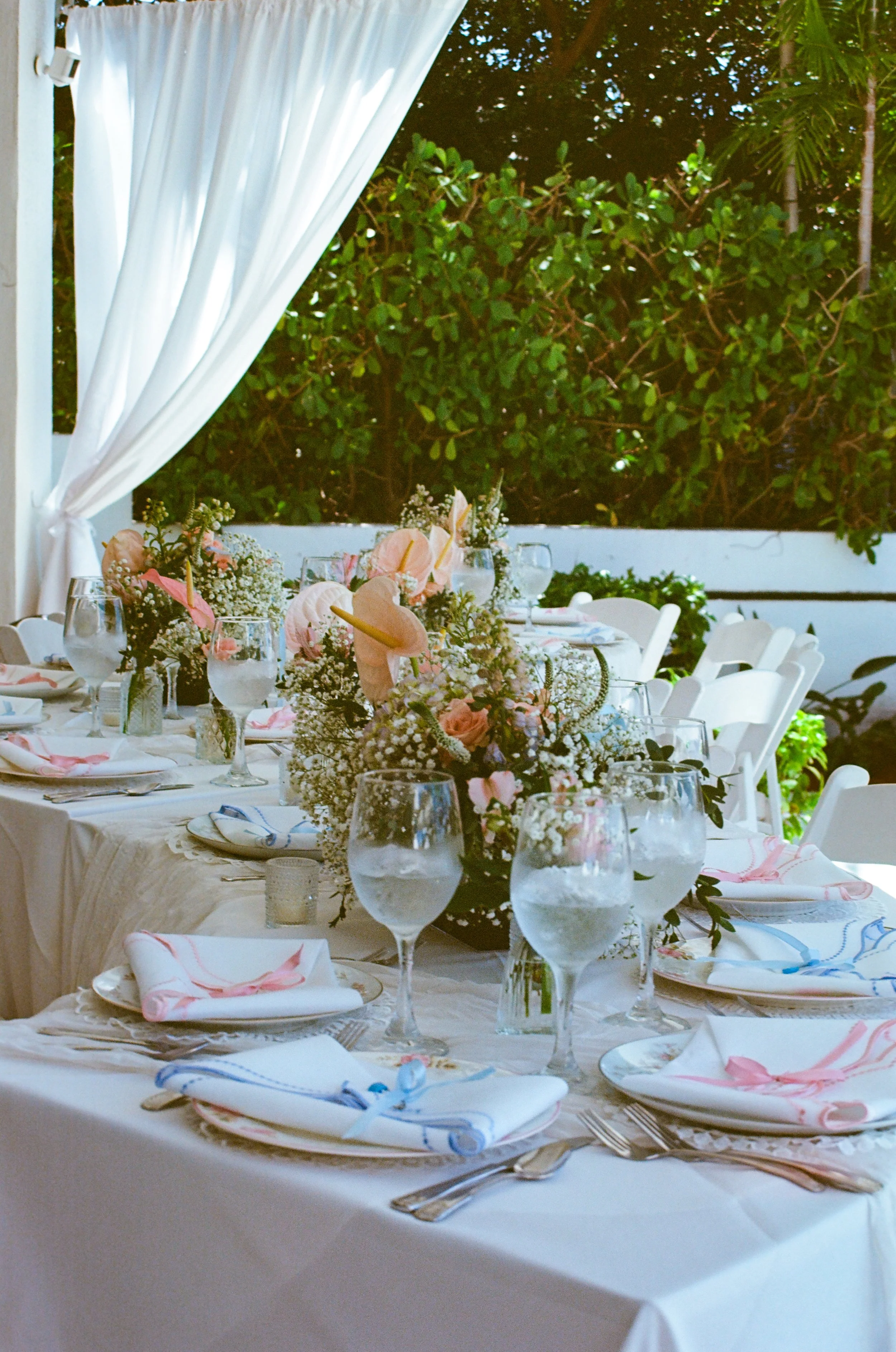 A beautifully set outdoor dinner table decorated with floral arrangements, wine glasses, plates with napkins tied with pink and blue ribbons, and silverware, with a white curtain on one side and lush green foliage in the background.