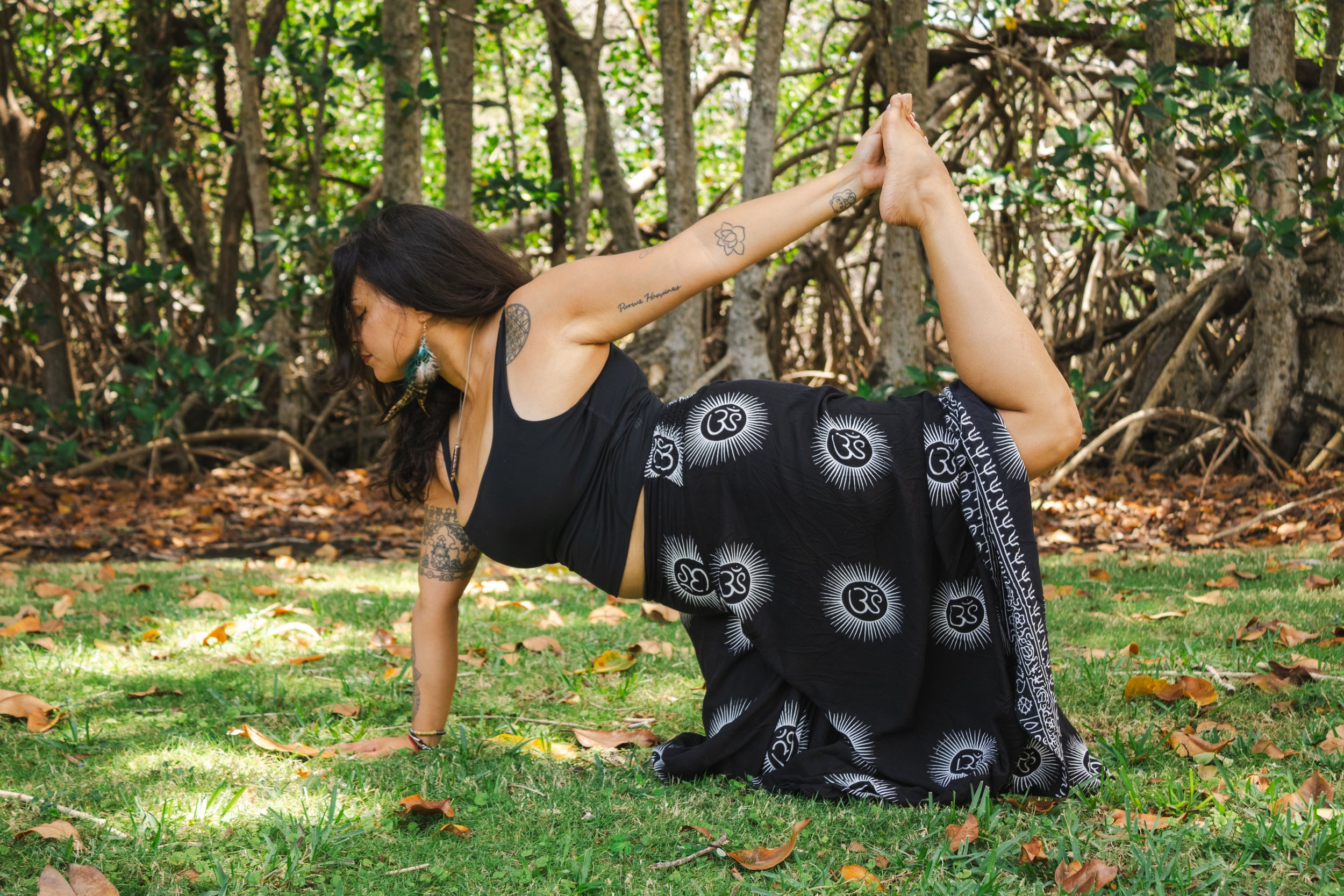 A woman practicing yoga outdoors in a forest, balancing on her hands with her legs bent and feet touching above her back, surrounded by trees and fallen leaves.
