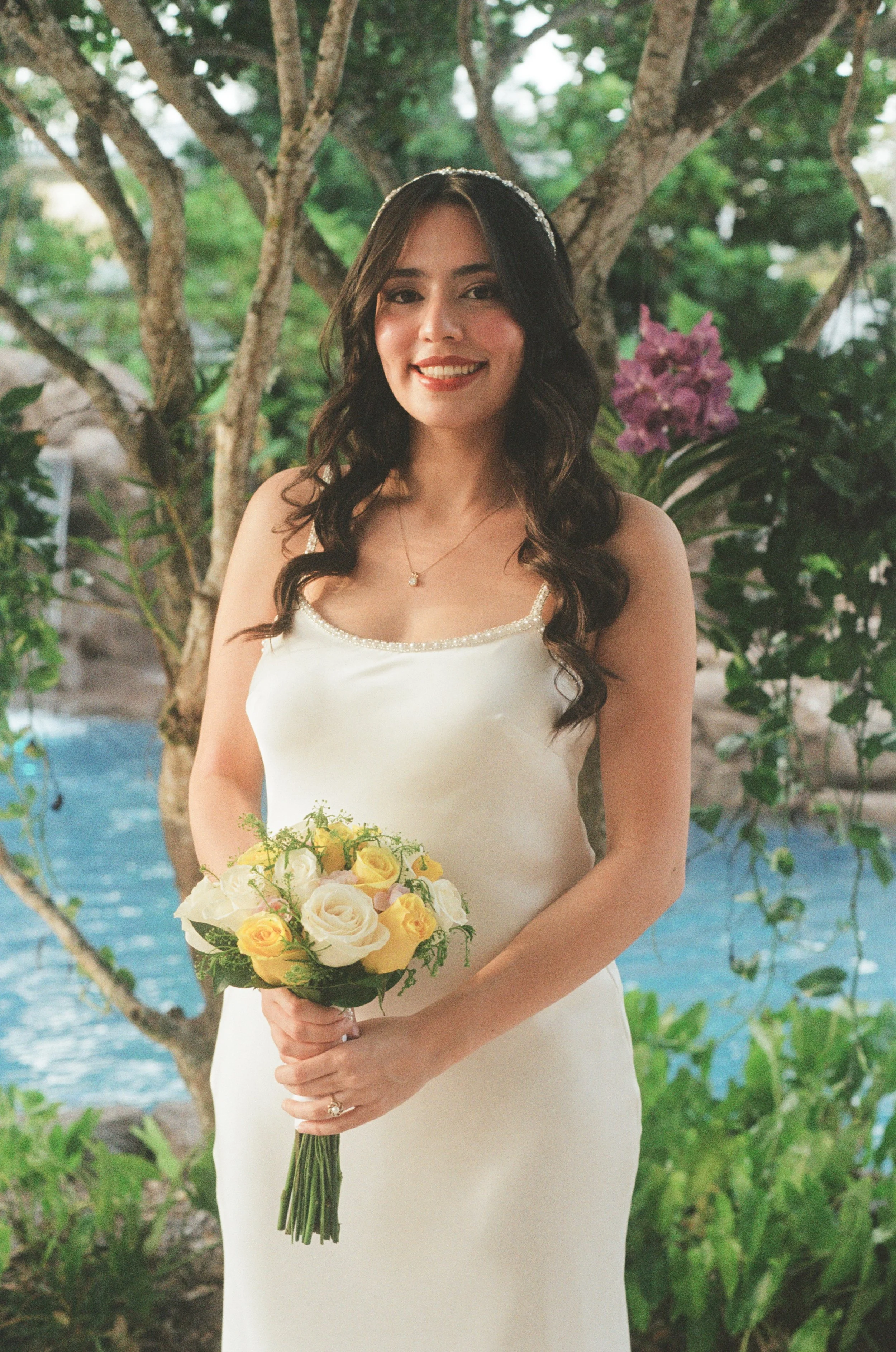 A woman in a white dress holding a bouquet of yellow and white roses, standing outdoors with green trees and purple flowers in the background.