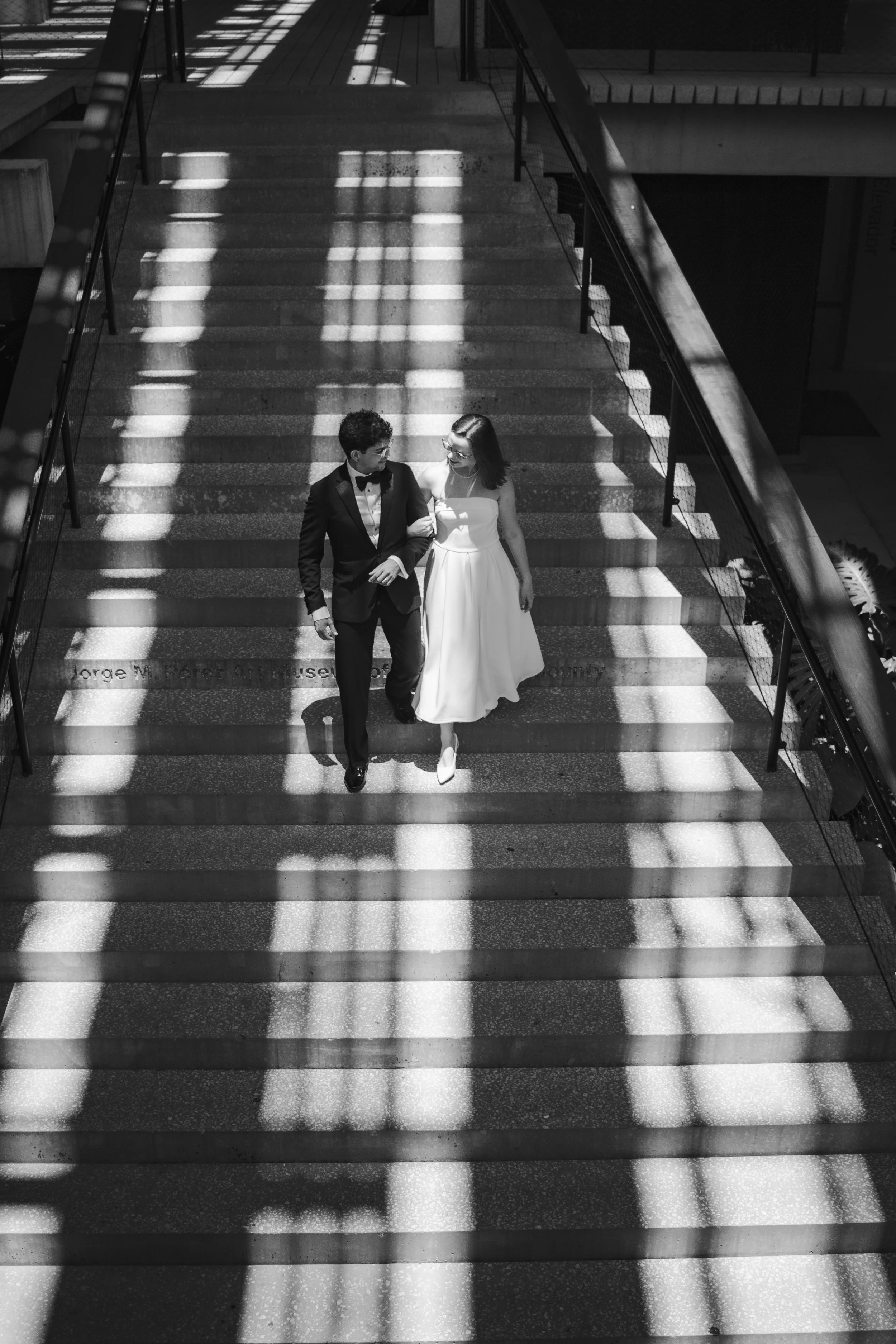 Black and white photo of a man and woman walking down stairs, with striped shadows cast on the stairs from the overhead structure.