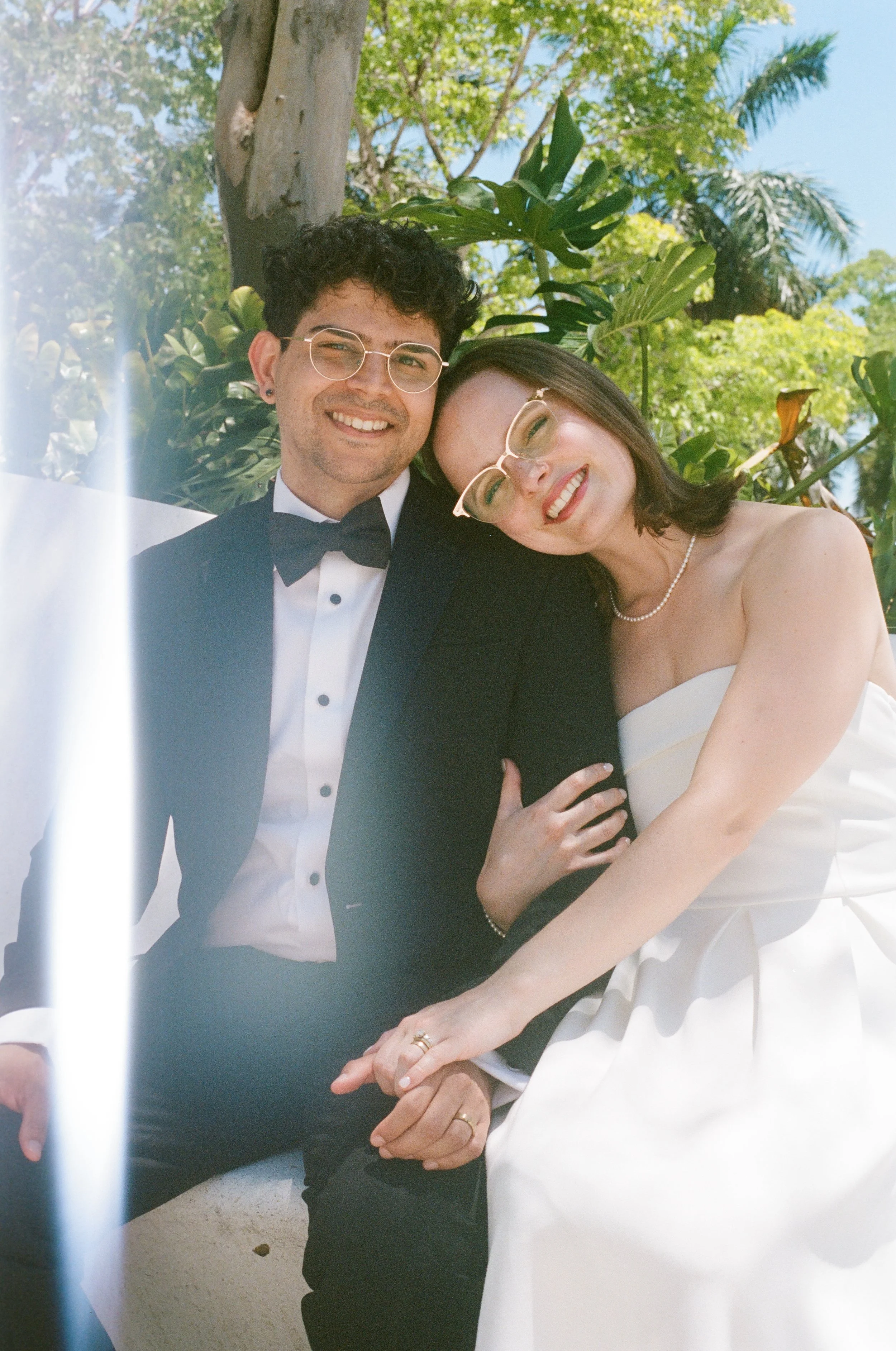 A smiling couple at a wedding, dressed in formal wedding attire, holding hands, with lush green trees and blue sky in the background.