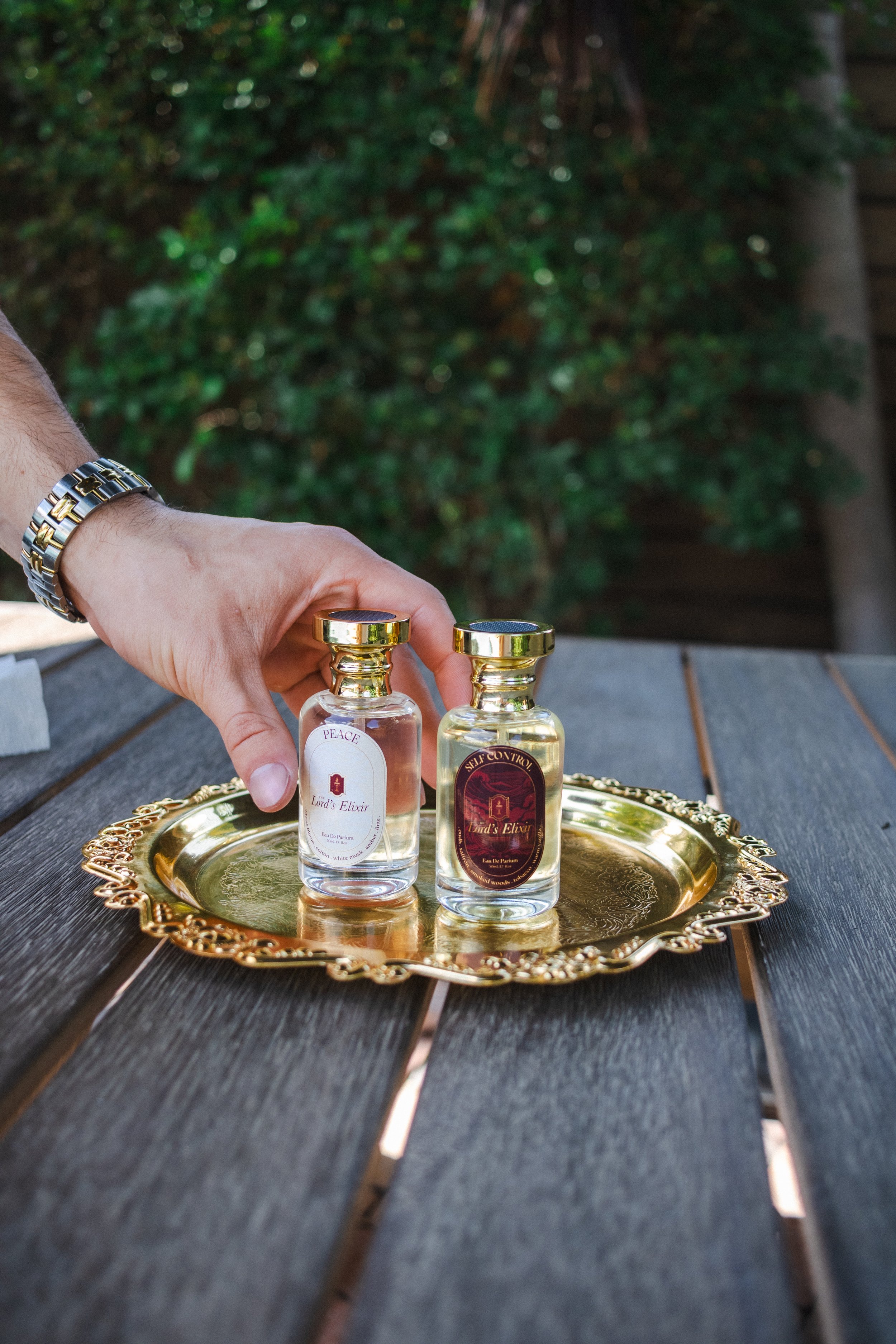 A person's hand, wearing a metal and gold watch, is reaching for two bottles of Lord's Elixir perfume on a gold decorative tray. The tray is on a wooden outdoor table with greenery in the background.