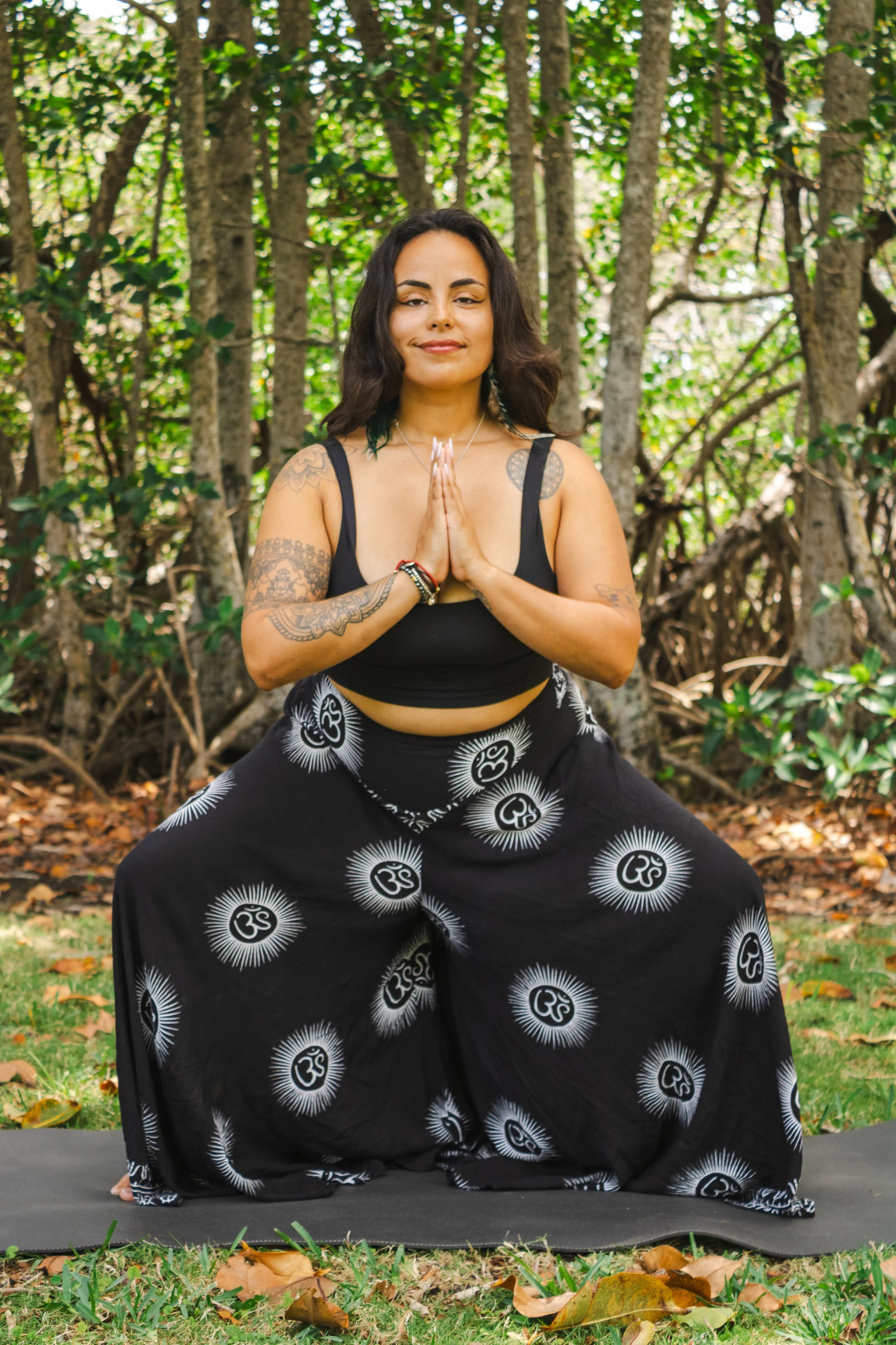 A woman practicing yoga outdoors on a black yoga mat, sitting in a wide-legged seated position with hands in prayer pose, surrounded by trees and fallen leaves.