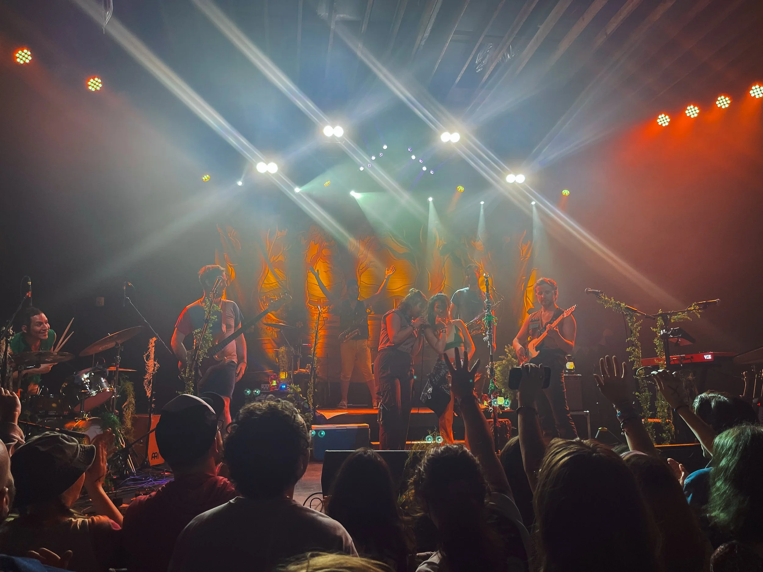 People performing live music on stage with bright stage lights and fog effects, audience watching in a darkened room.