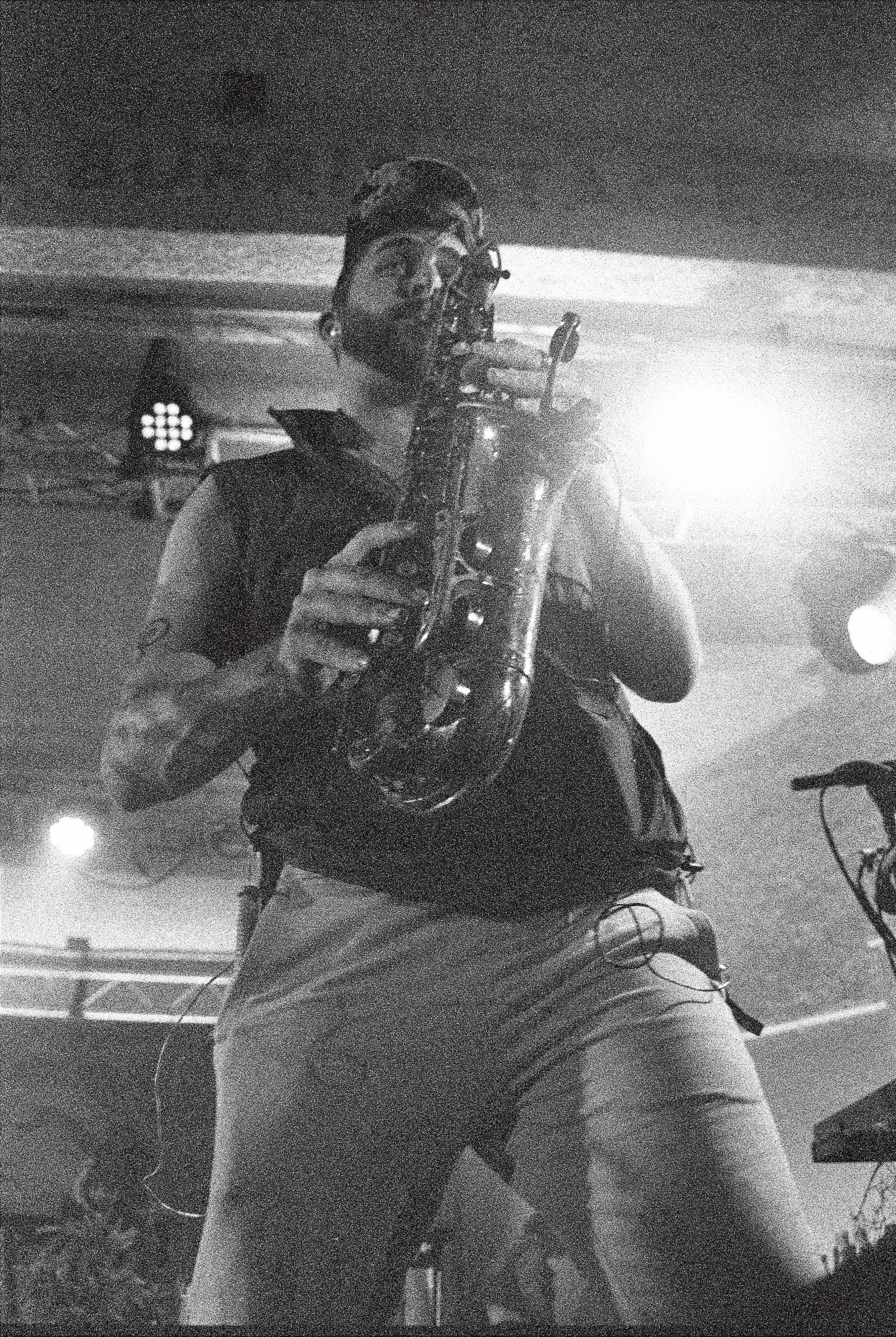 A person with long hair, glasses, and headphones, holding a saxophone in a black-and-white photo.