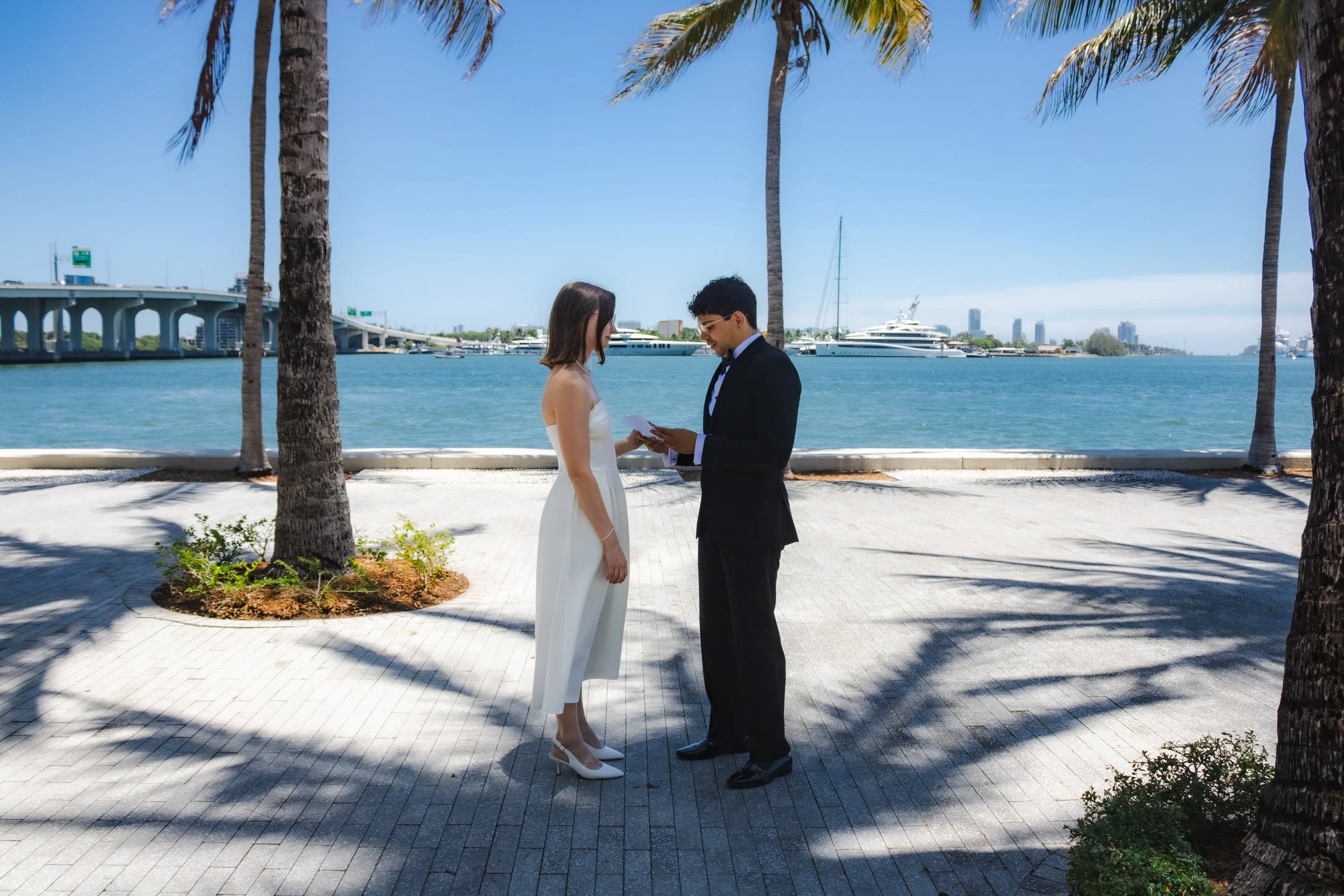 A couple dressed for a wedding, standing face to face near a waterfront with palm trees, a bridge, and yachts in the background. The bride is in a white wedding dress, and the groom is in a dark suit with a tie.