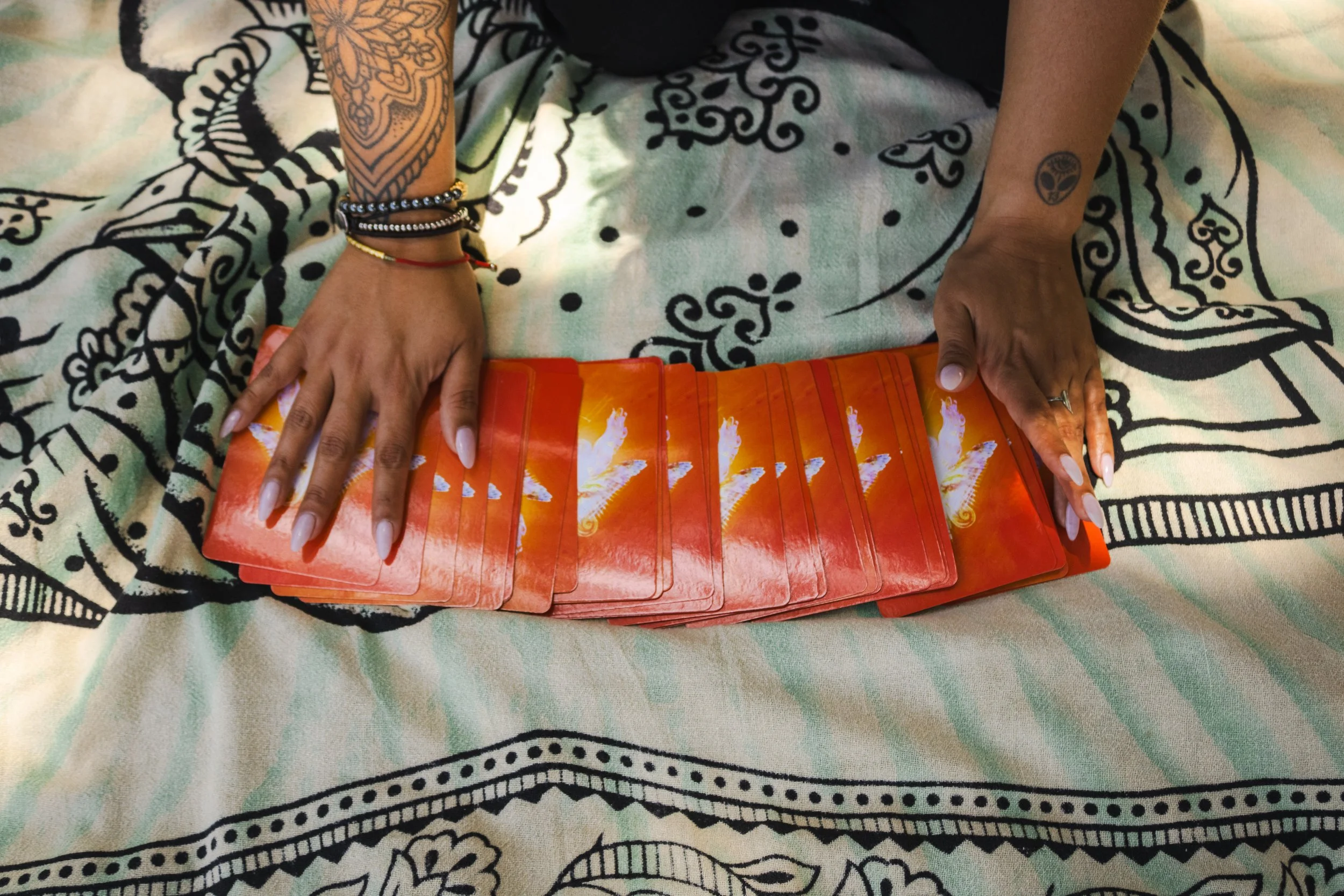 Person with tattoos on both arms and multiple bracelets, holding a deck of red and orange tarot cards on a black and white patterned cloth.
