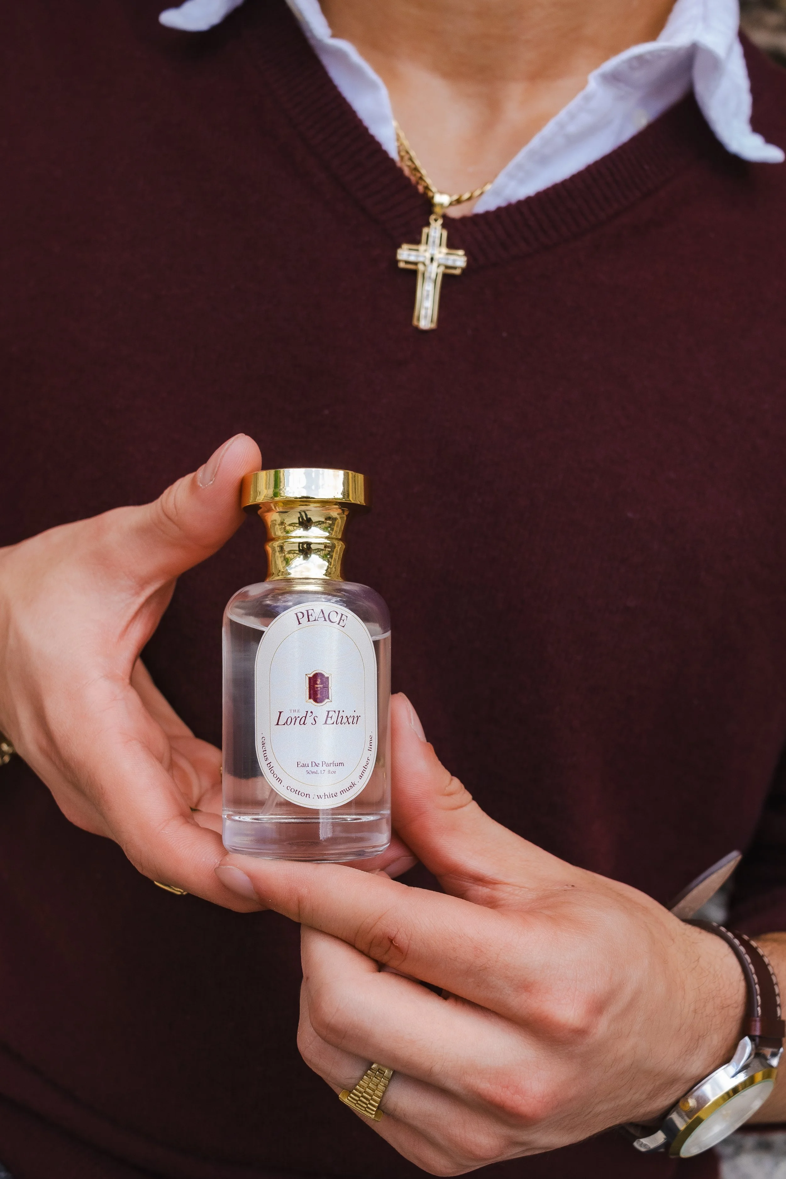 Person holding a small perfume bottle labeled 'The Lord's Elixir' and wearing a gold cross necklace over a maroon sweater with a white shirt underneath.