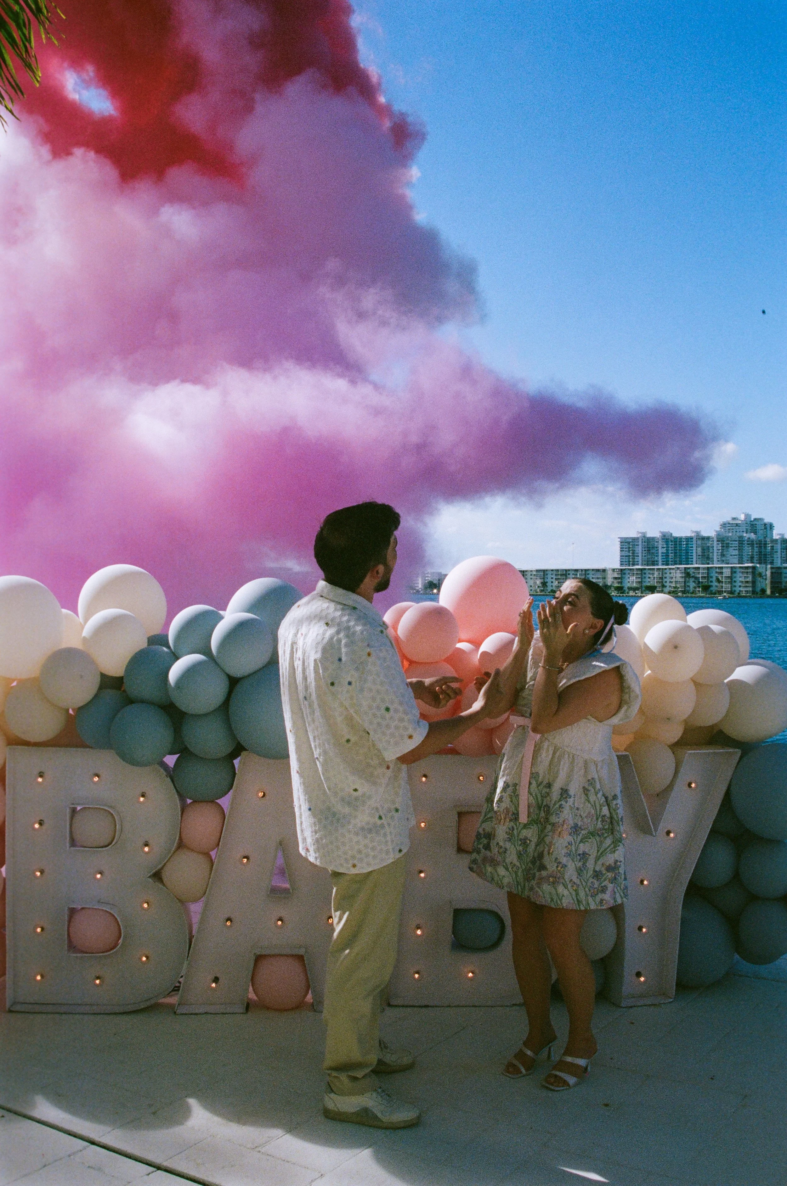 A man and woman are talking in front of a large sign that says 'BAR' with balloons attached. The sky is filled with purple and pink clouds, and there are city buildings in the background.