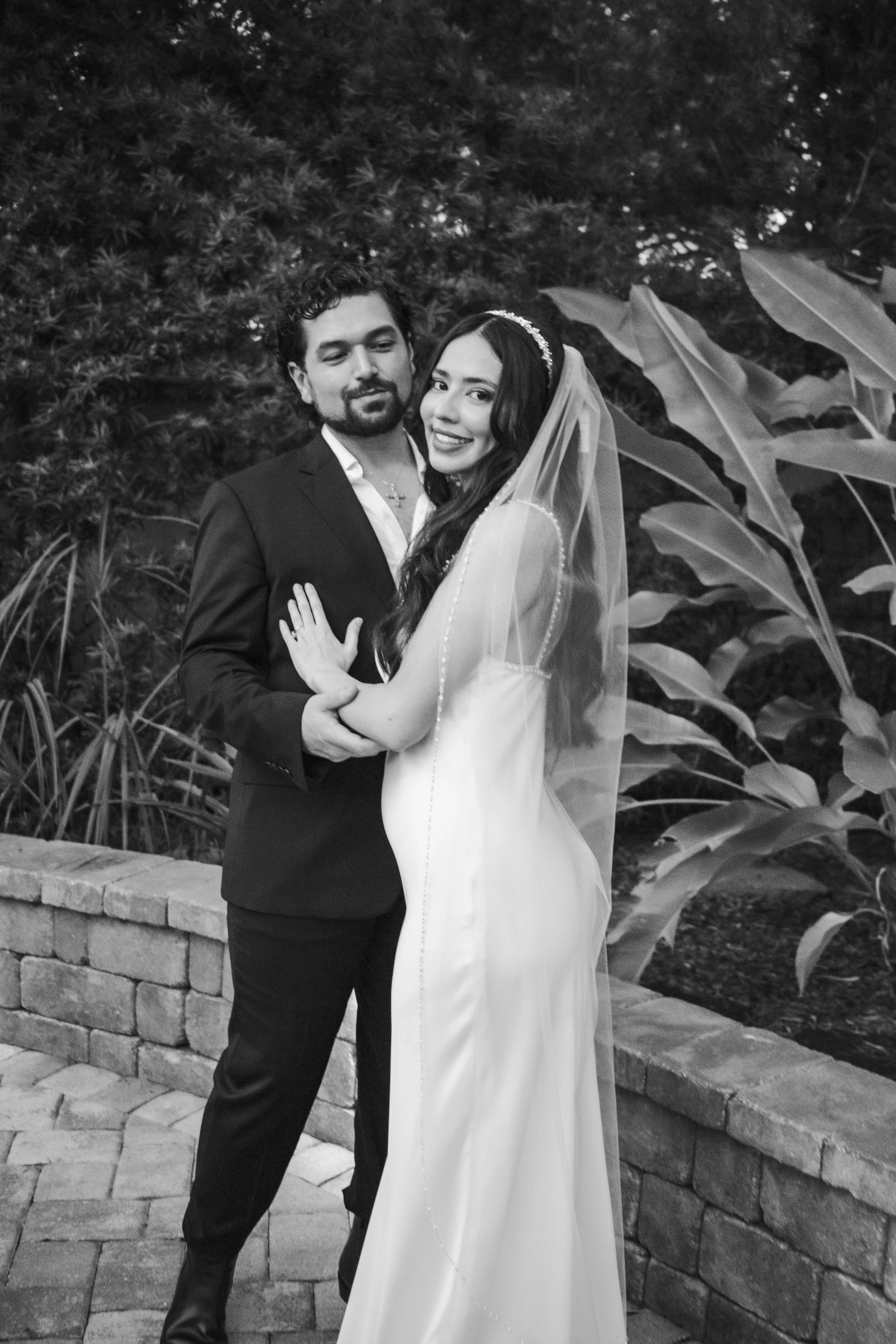 A black-and-white photo of a bride and groom standing close together outdoor, with large leafy plants in the background. The groom is wearing a suit and the bride is wearing a wedding dress with a veil and headband. They are smiling and holding each 