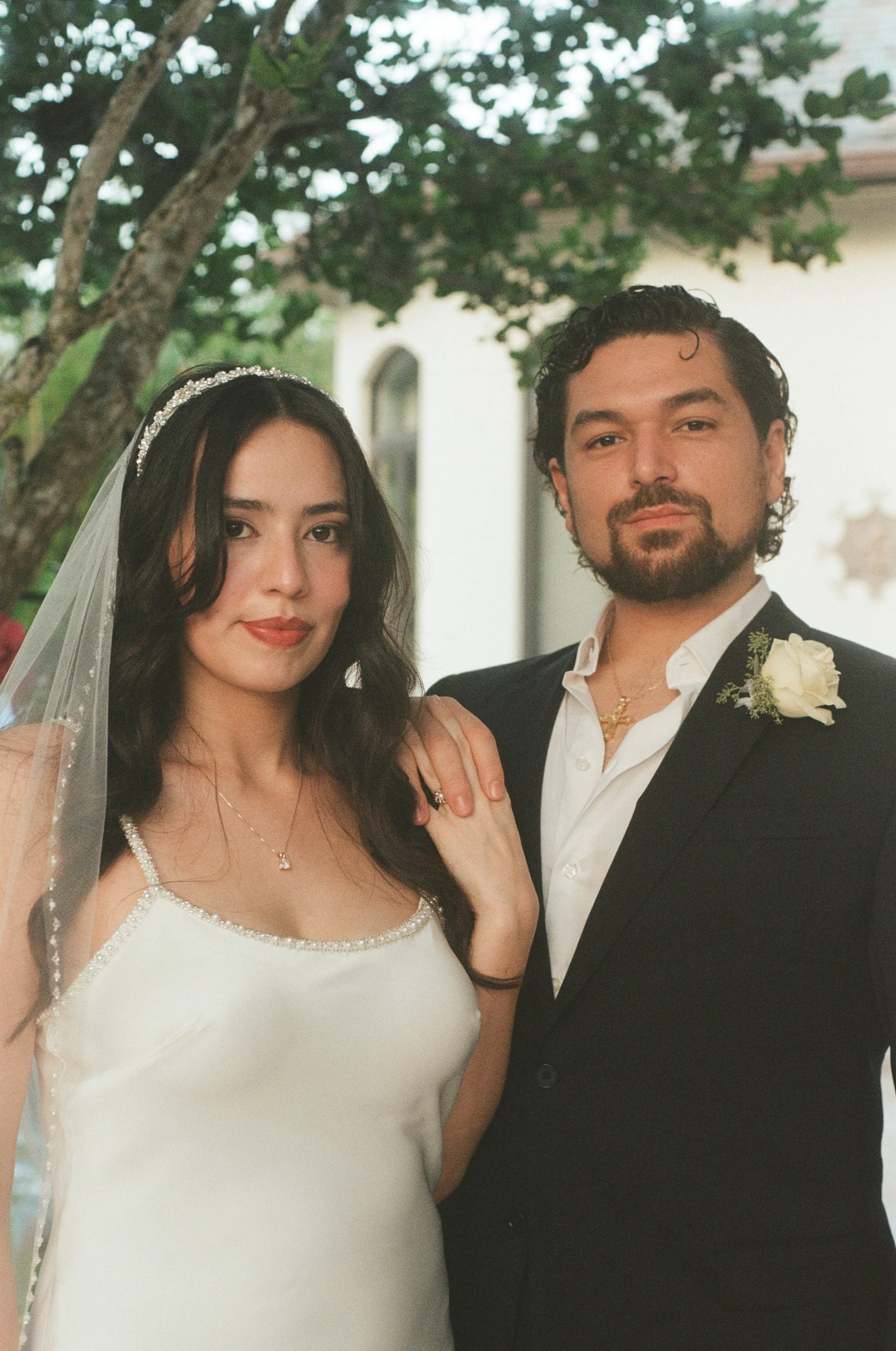 A bride and groom dress formally for their wedding, standing outdoors under a tree. The bride is wearing a white wedding dress with beaded accents and a veil, and the groom is dressed in a black tuxedo with a white shirt and boutonniere. They are loo