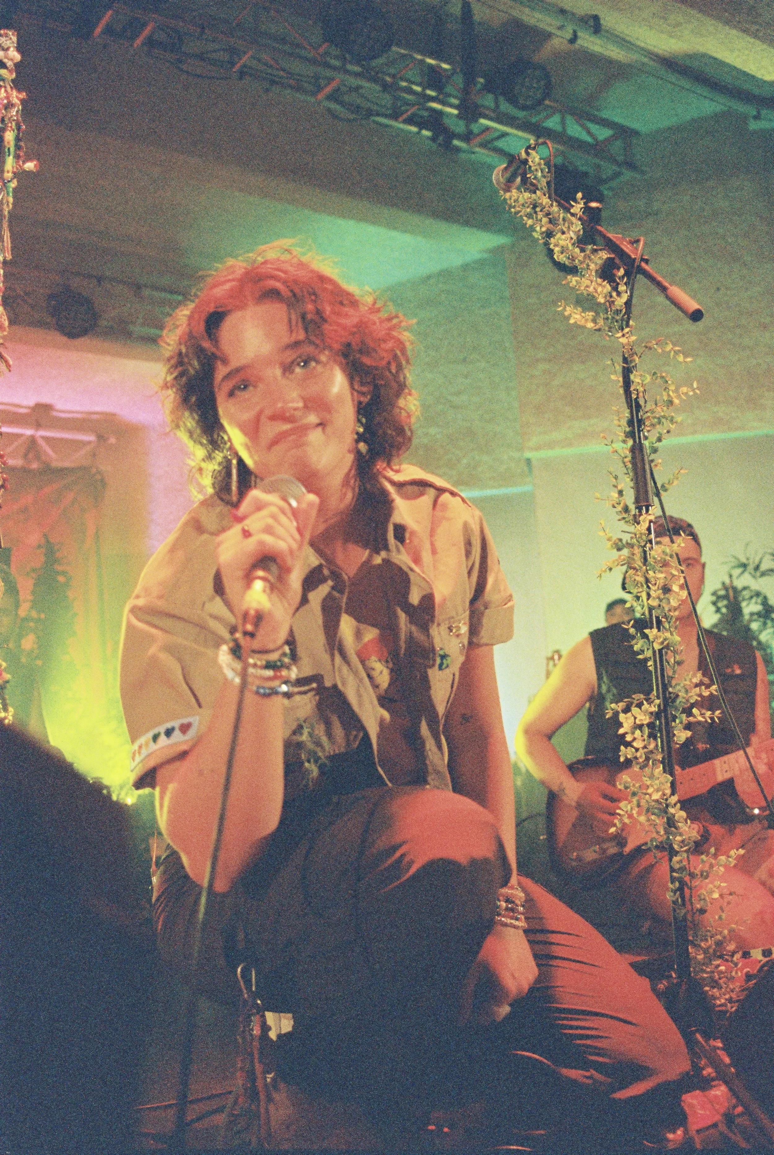 A woman with curly hair holding a microphone, sitting on a stage with colorful lighting, surrounded by microphone stands and plants, with other musicians in the background.