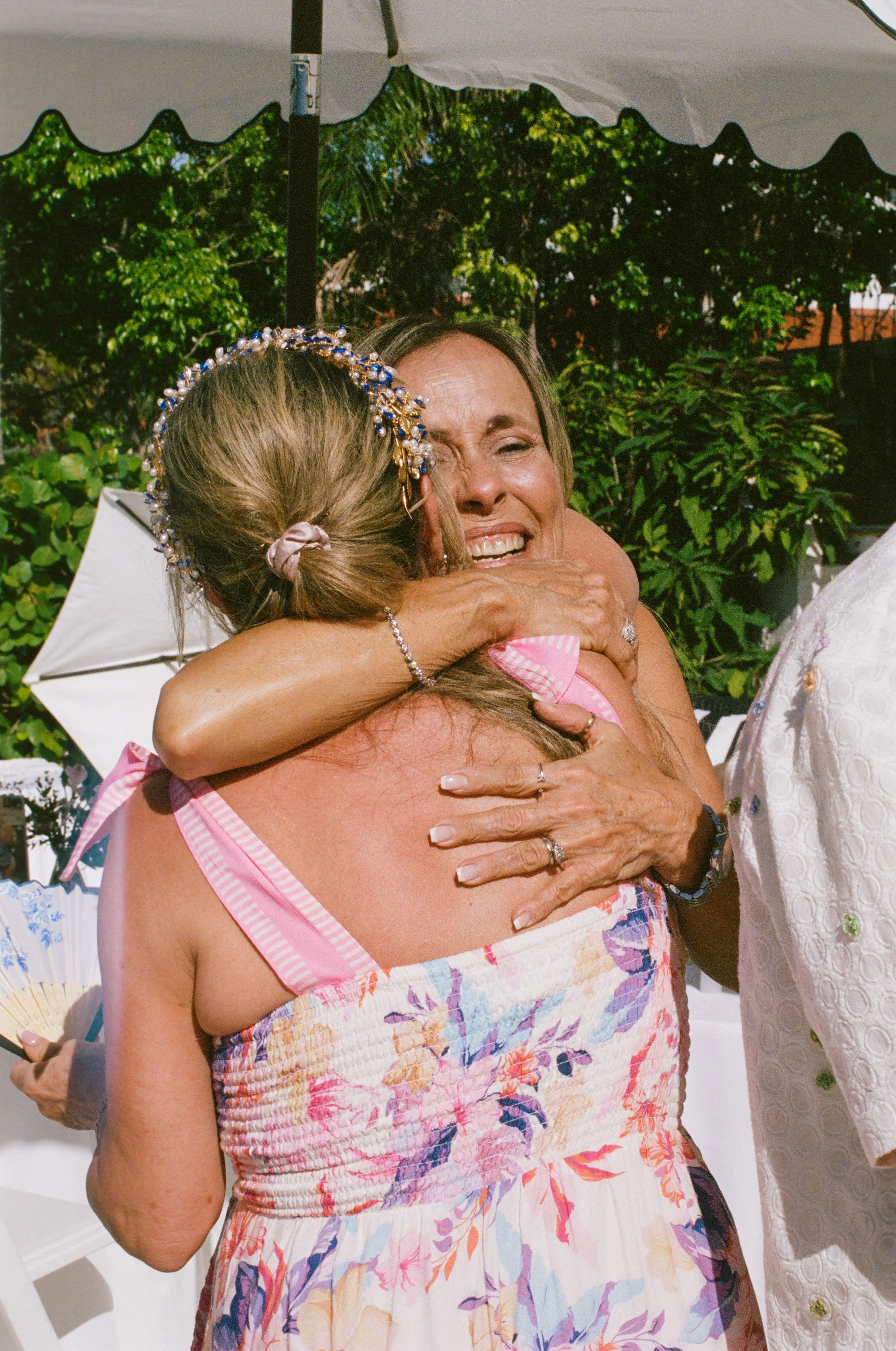 Two women hugging and smiling at an outdoor event, surrounded by greenery under a tent.