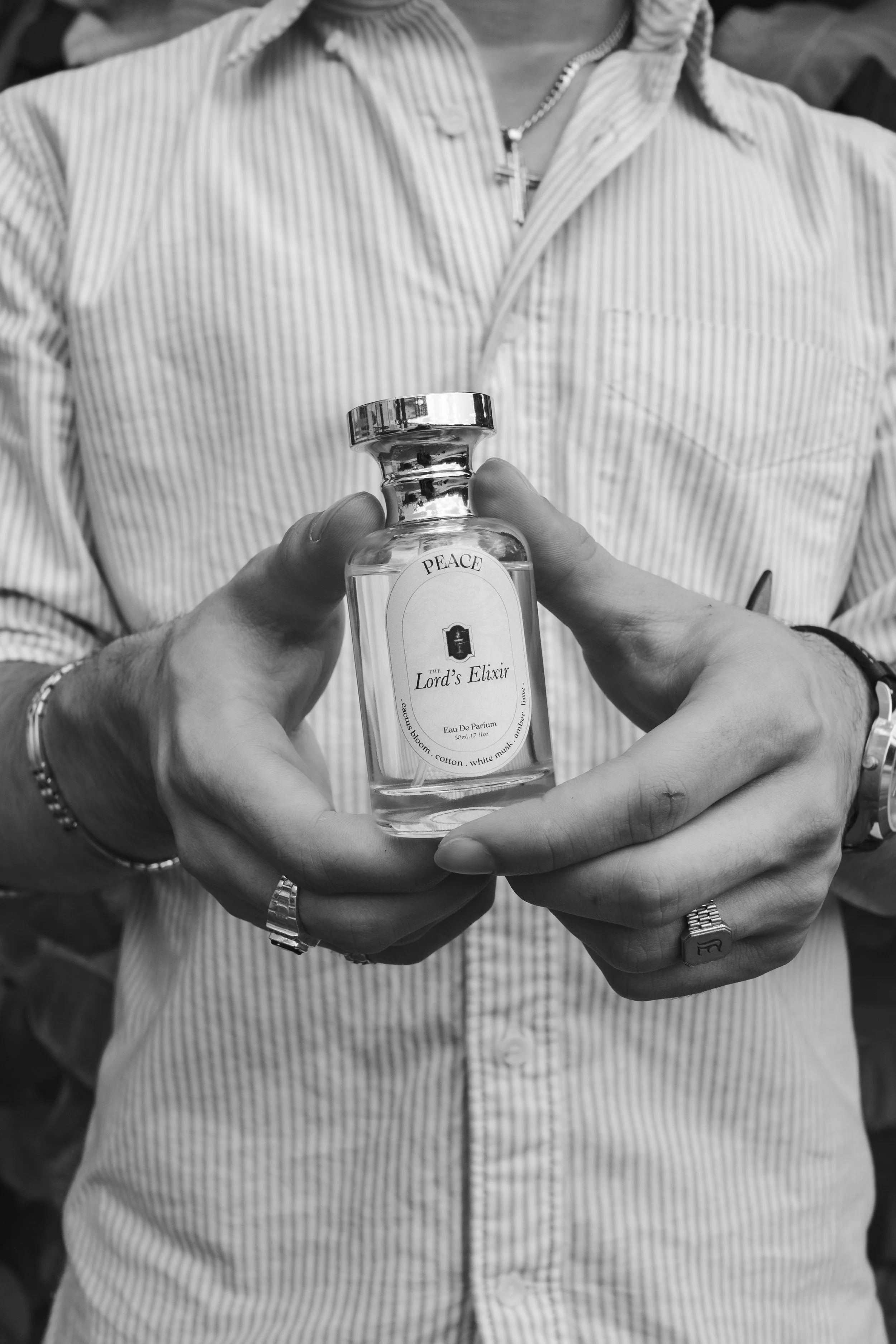 Person wearing a striped shirt and jewelry holding a small bottle labeled 'Lord's Elixir' with the word 'PEACE' on top.