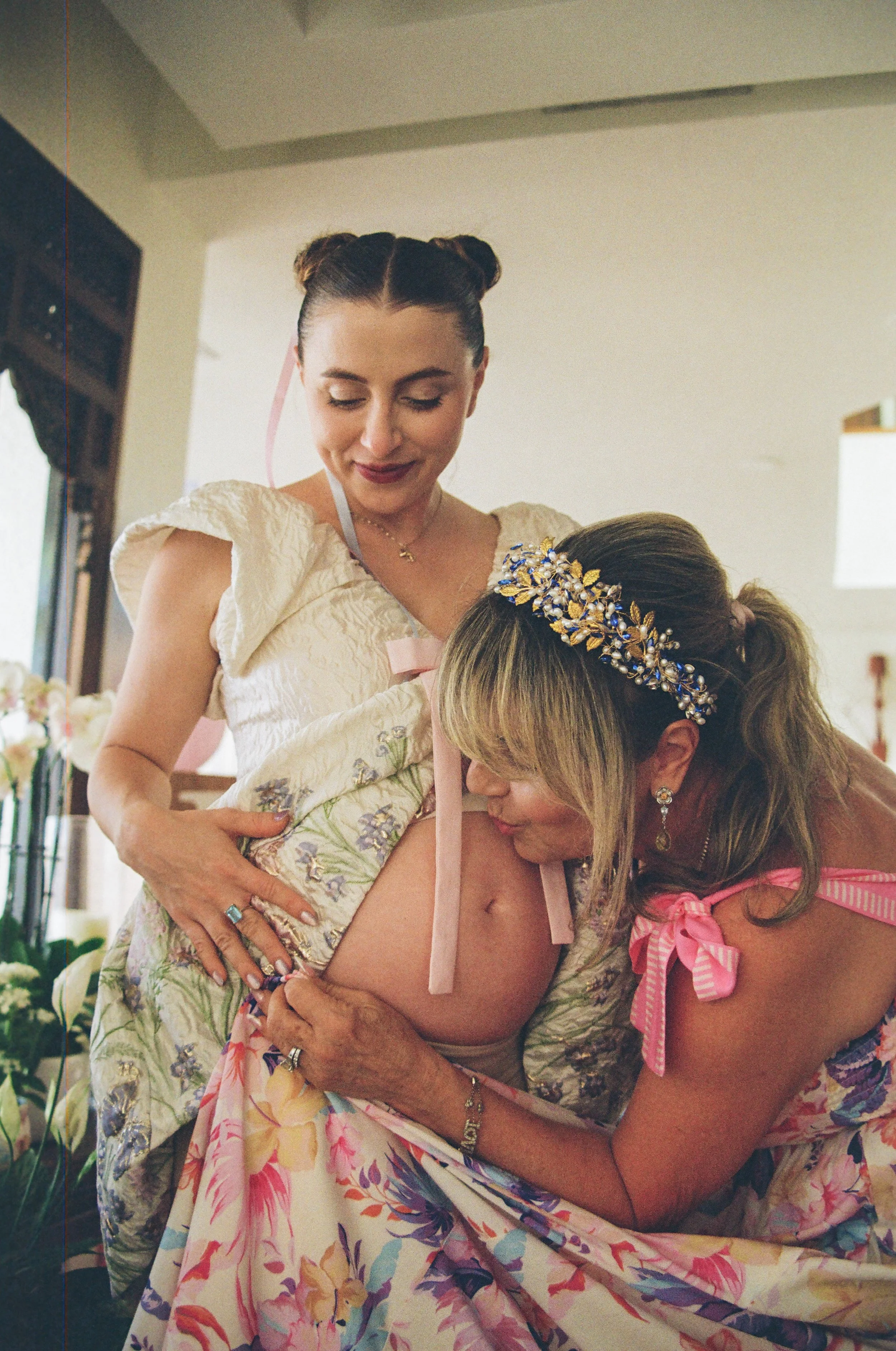 A woman with styled brown hair and a woman with blonde hair are embracing each other. The woman with styled brown hair is wearing a floral dress with puffed sleeves and a gold necklace. The woman with blonde hair is wearing a floral dress with pink r