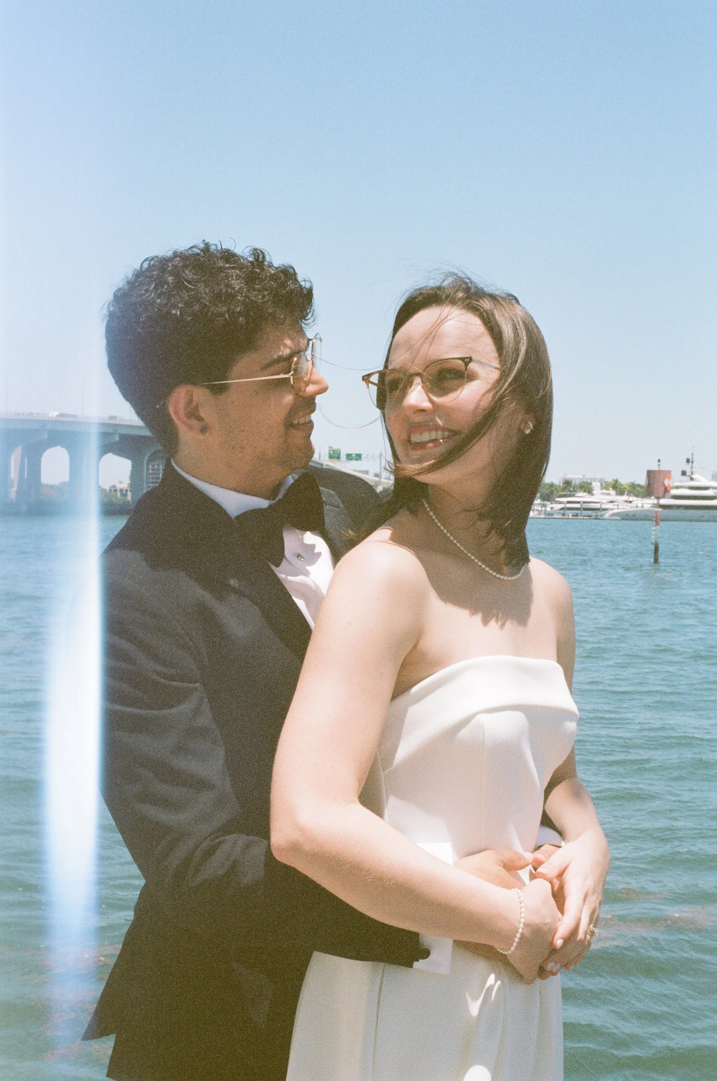 A couple dressed in wedding attire, the man in a tuxedo and the woman in a white dress and sunglasses, standing by the water with boats and a bridge in the background.