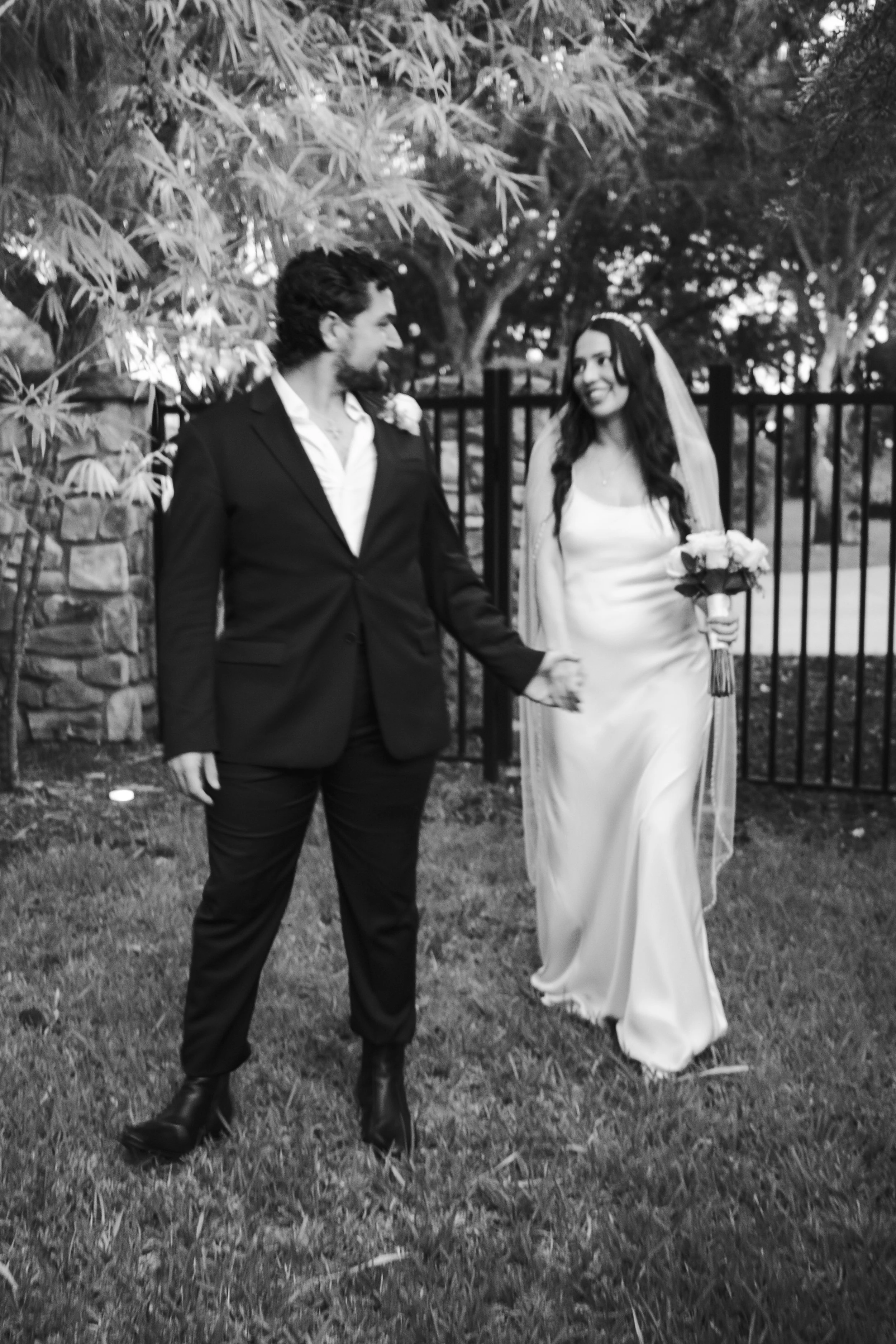 A black and white photo of a couple at a wedding, holding hands, with a fence and trees in the background. The man is wearing a suit and the woman is in a wedding dress with a veil and is holding a bouquet of flowers.