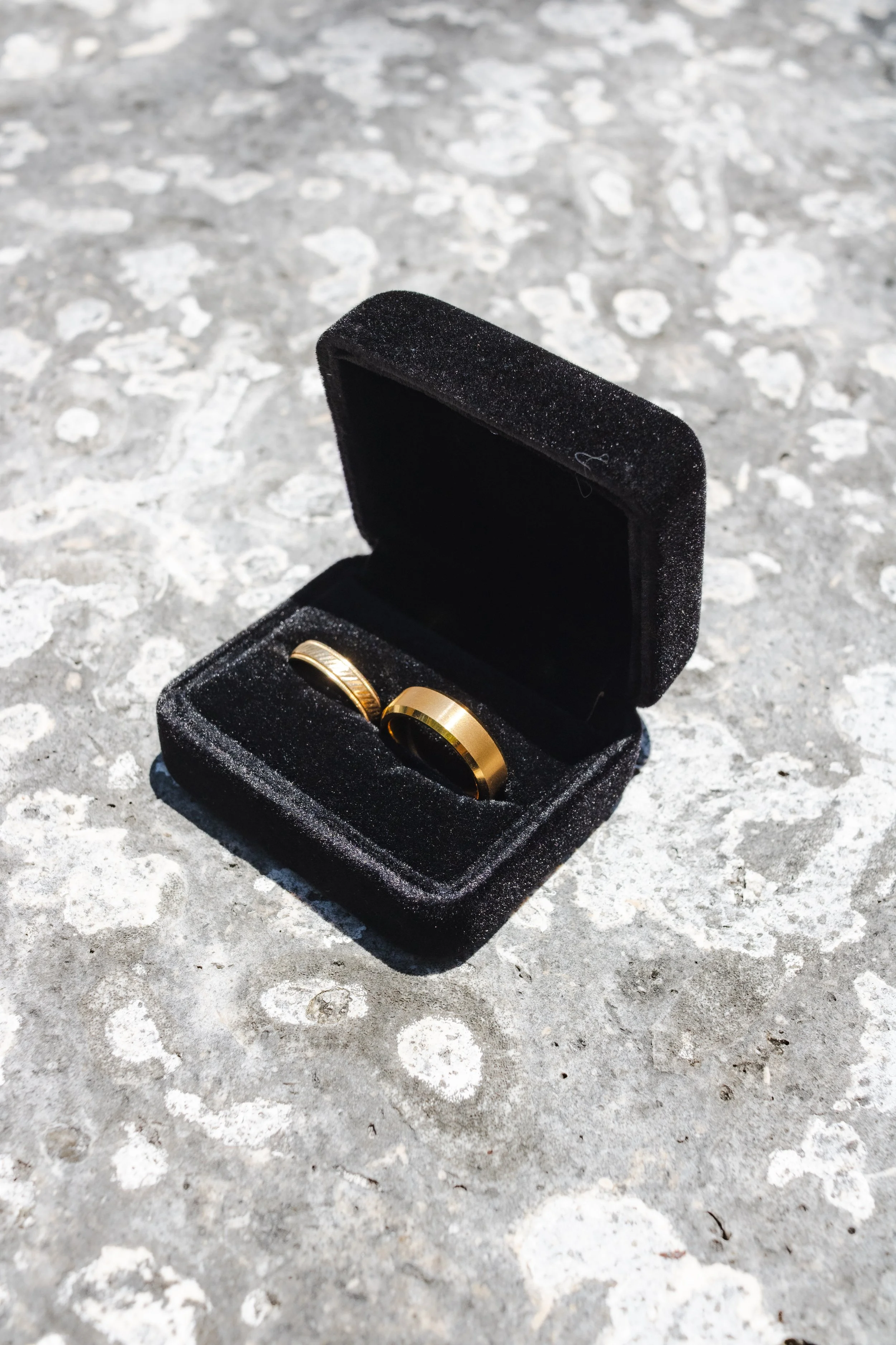 A small black jewelry box with a velvet-like texture, open to reveal two gold wedding bands inside, placed on a rough, light-colored concrete surface.