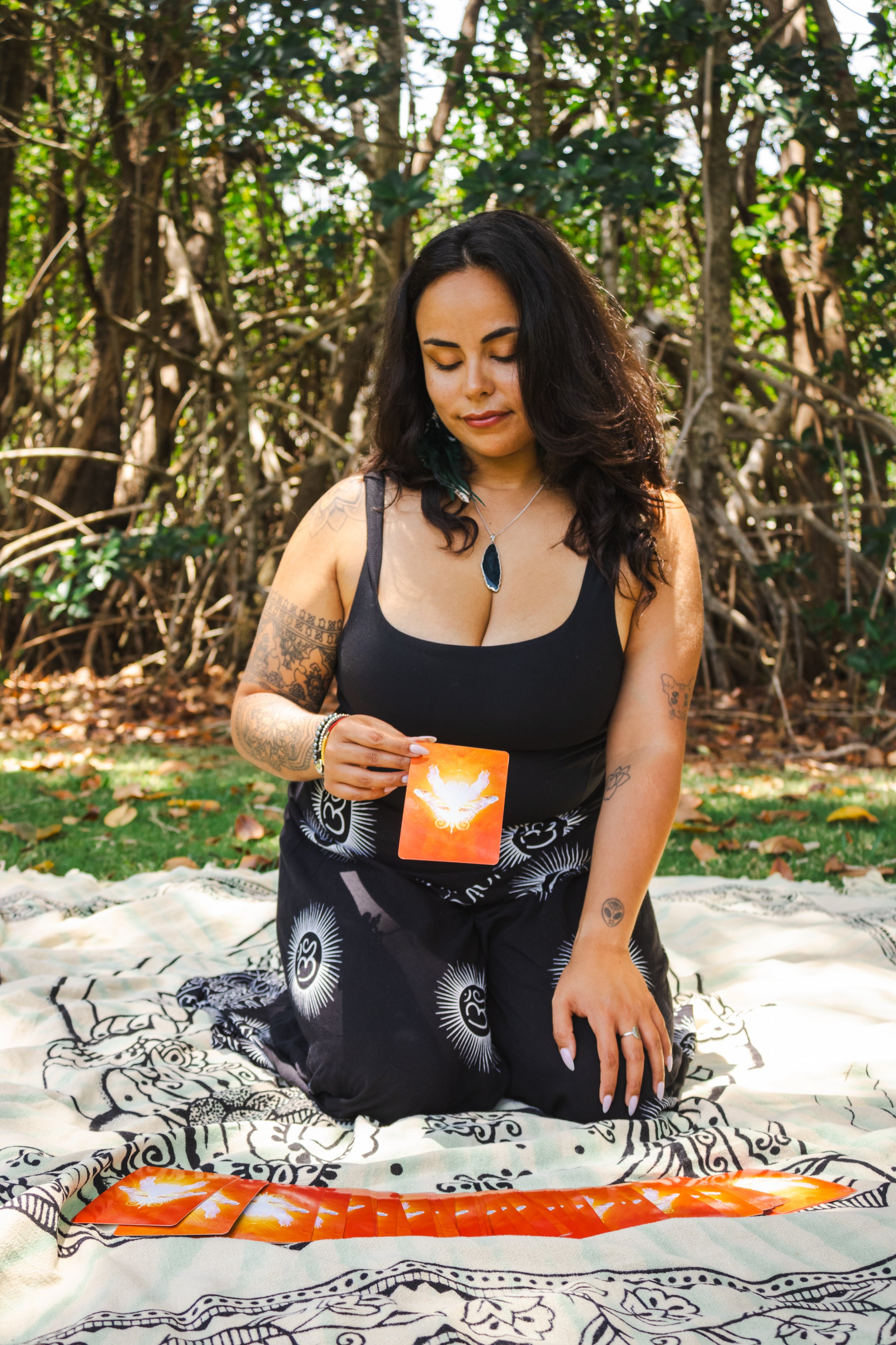 A woman with dark wavy hair and tattoos on her arm is kneeling on a patterned blanket outdoors in a forest setting. She is holding tarot cards and a tarot card with an angel on it is visible in front of her.