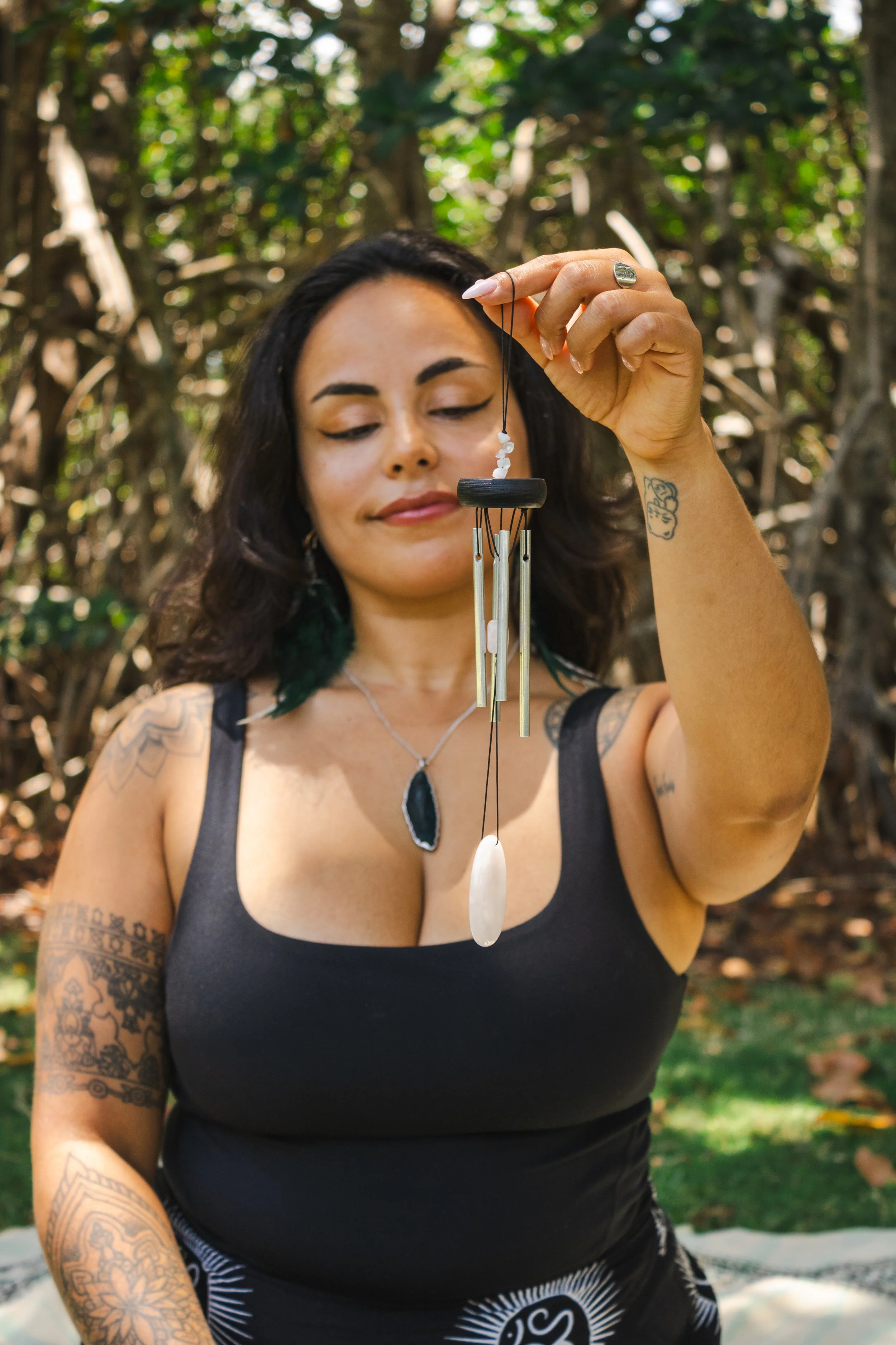 A woman with tattoos and dark hair, wearing a black tank top, holds a wind chime made of metal and shells outdoors with trees and greenery in the background.
