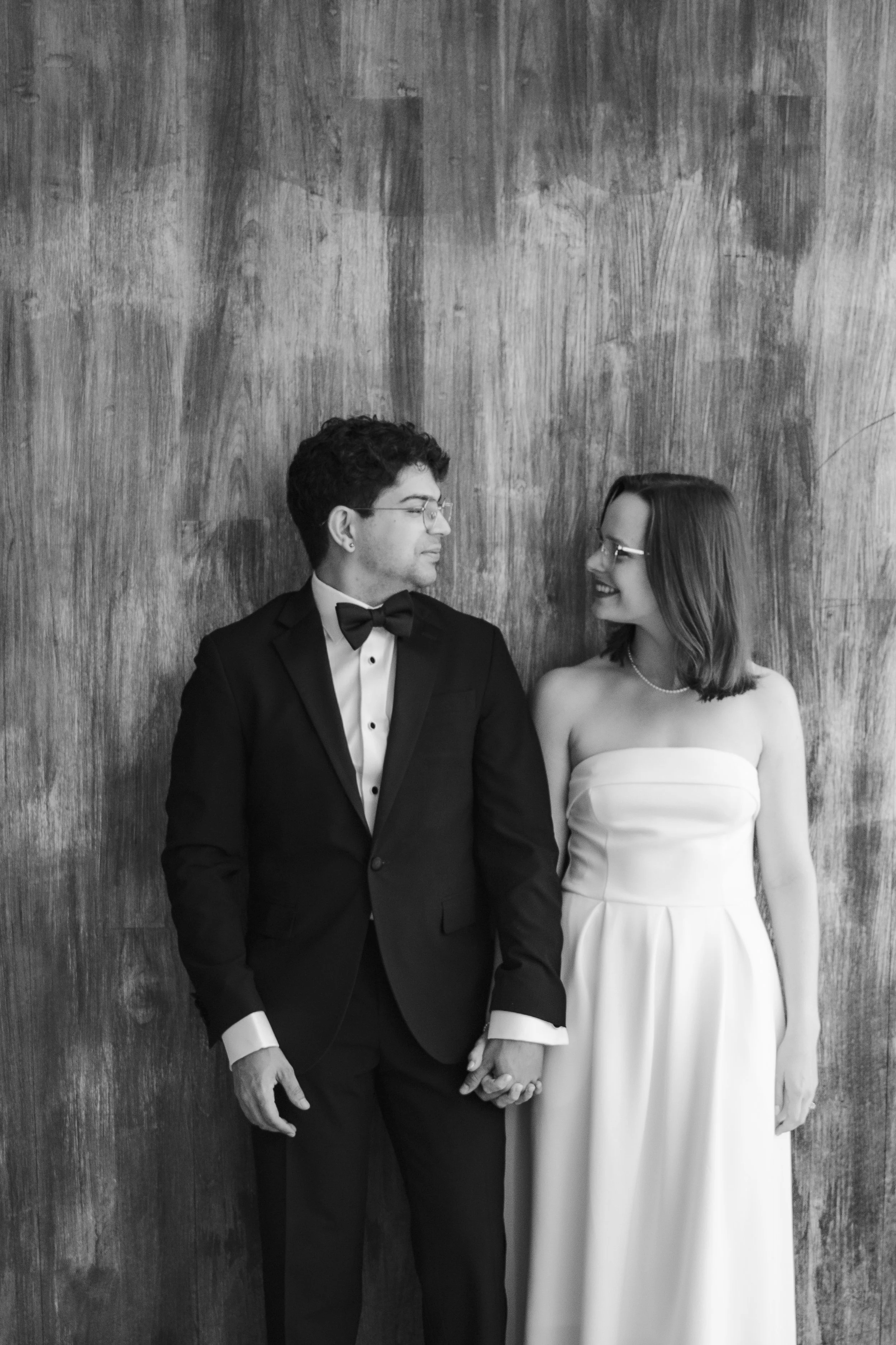 A black-and-white photo of a man and woman dressed in formal wedding attire, standing against a wooden wall. They are holding hands and looking at each other, smiling.