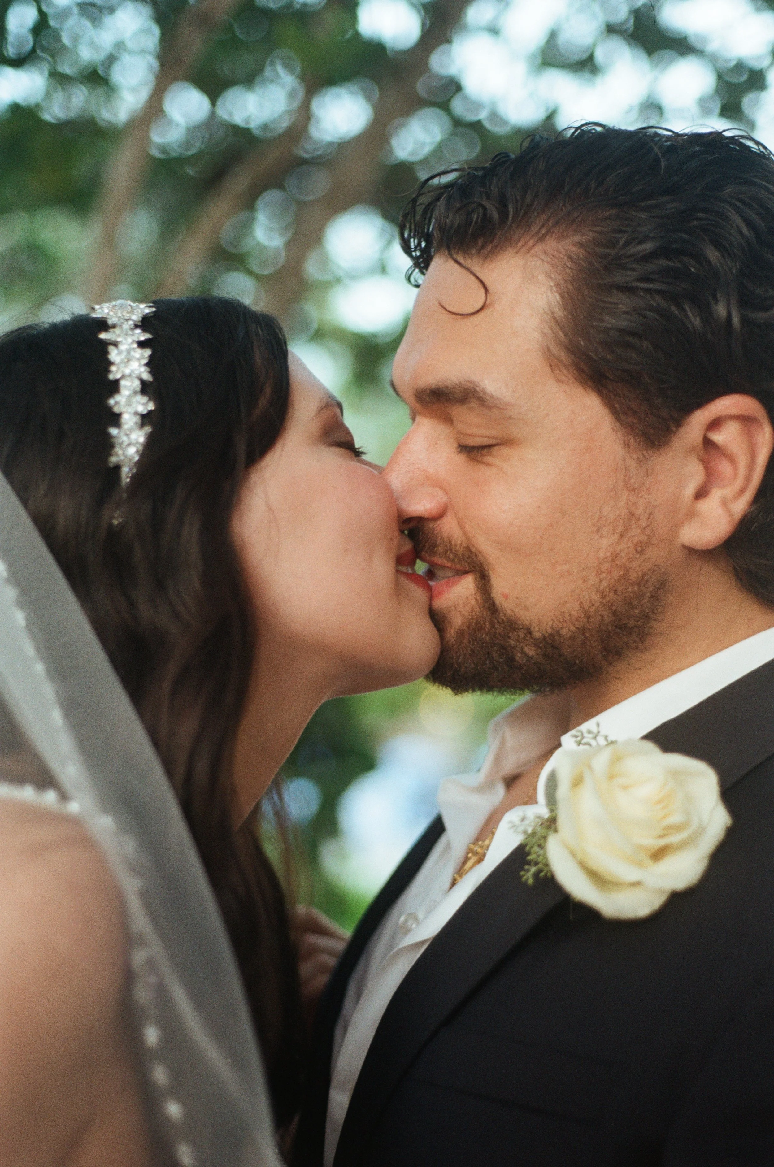 A bride and groom are sharing a kiss during their wedding ceremony outdoors, surrounded by soft bokeh lights and greenery.