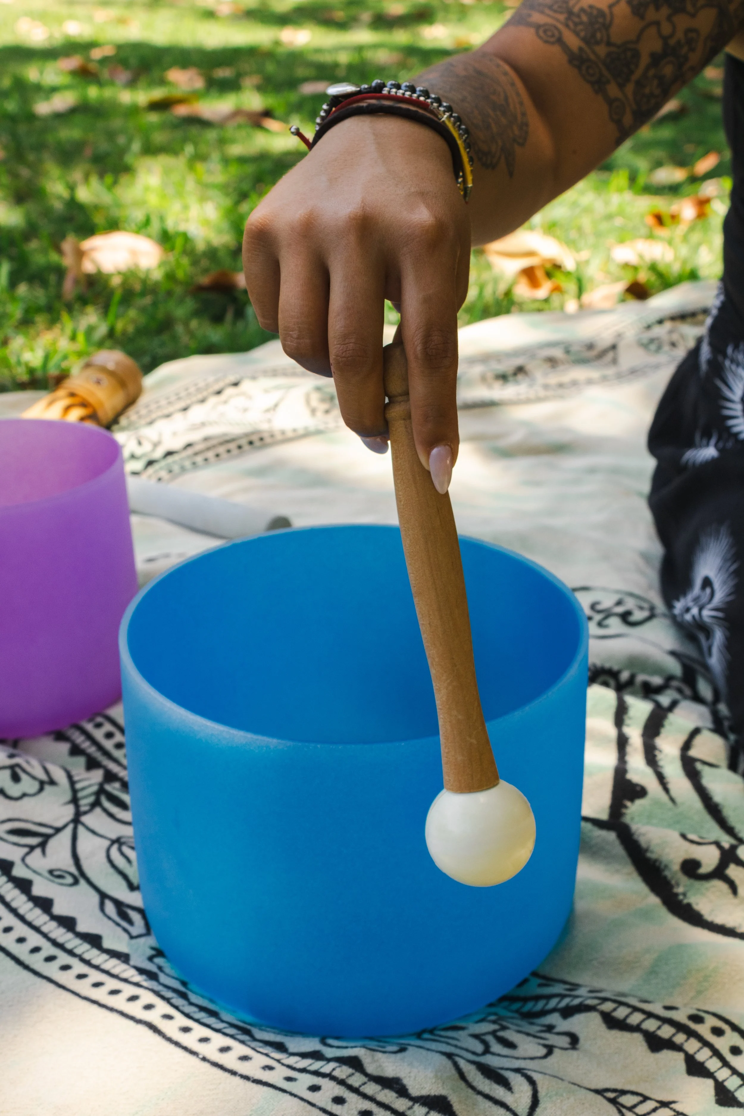 Close-up of a hand placing a wooden Mallet into a blue singing bowl on a patterned cloth outdoors, with another purple singing bowl nearby.