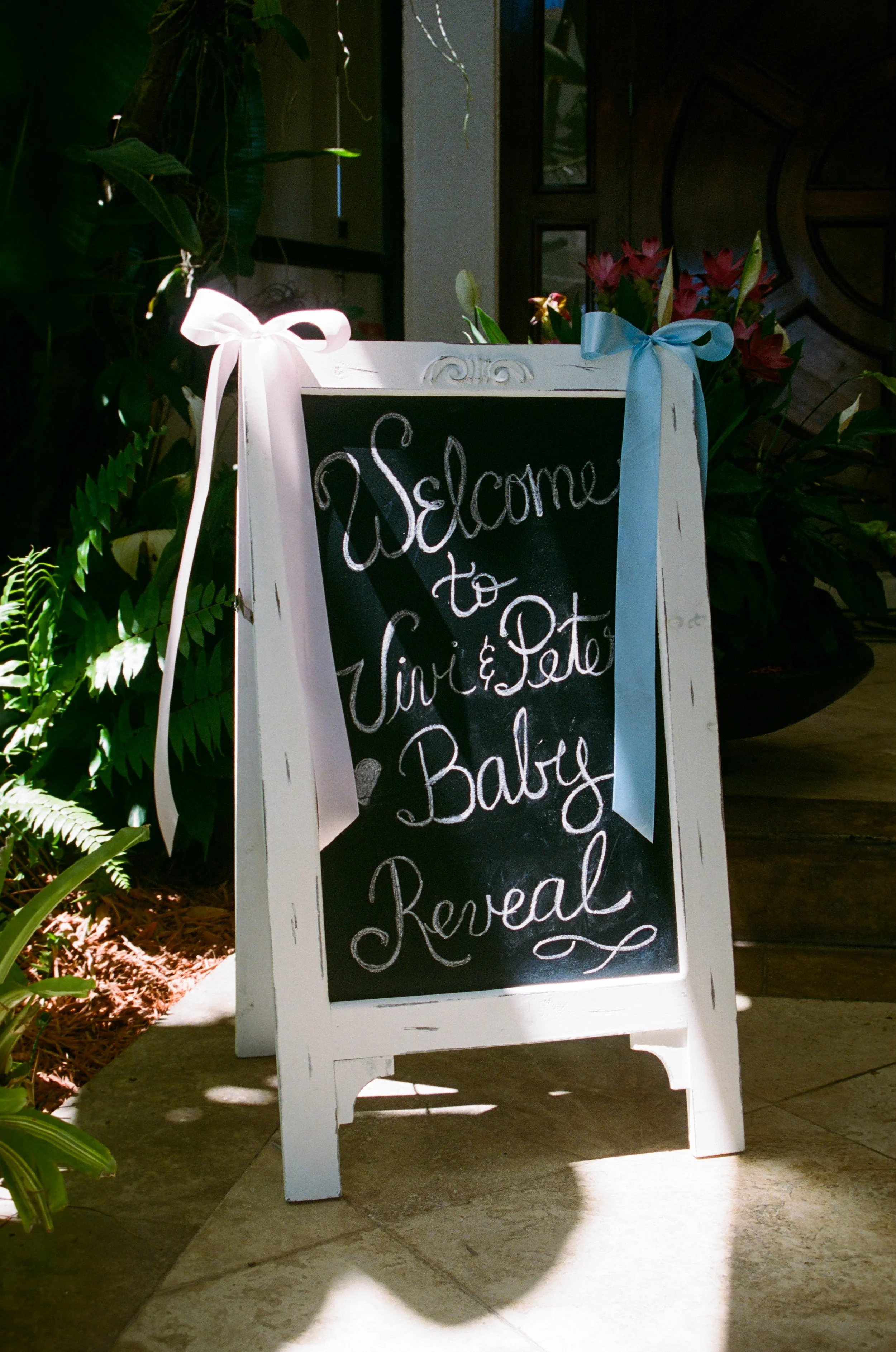 A white A-frame sidewalk sign with a chalkboard surface that reads "Welcome to Gin & Tonic & Balti" in white chalk, decorated with pastel ribbons, placed on a tile floor near green plants and a wall with a circular wooden decor piece.