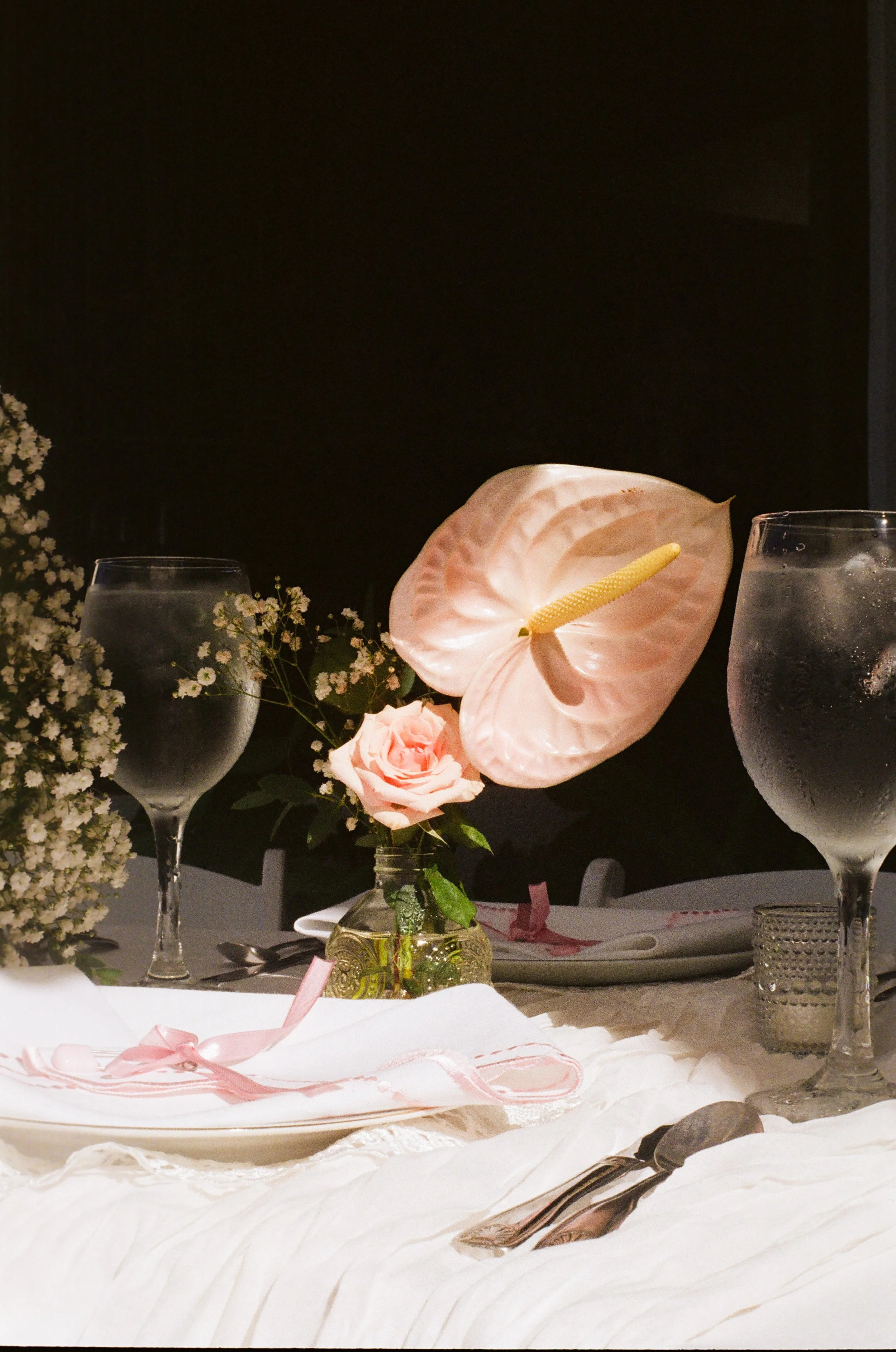 A table set for a celebration with pink and white flowers, wine glasses, and pink ribbons, against a dark background.