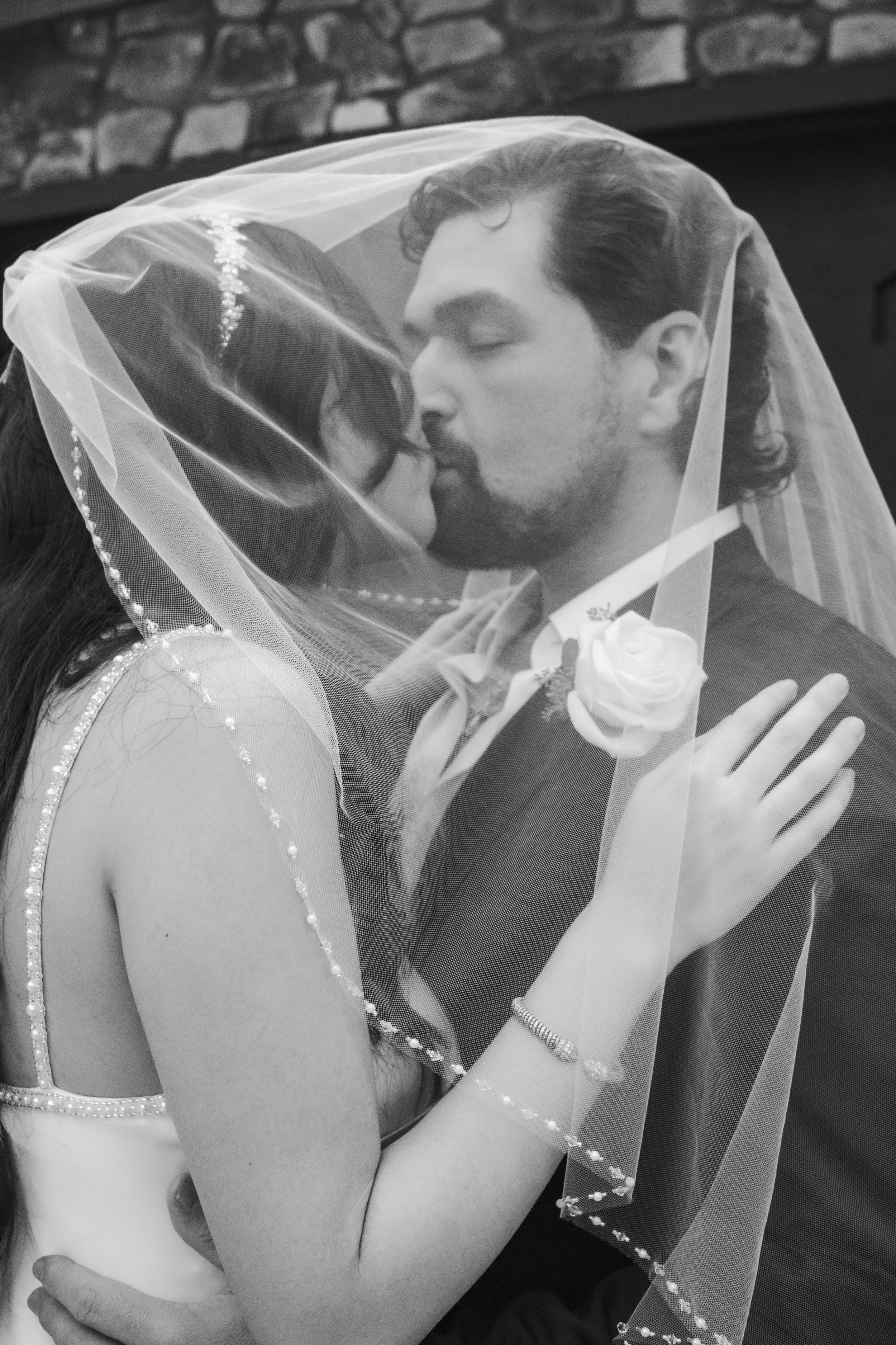 A black and white photo of a couple kissing, with a veil covering both of their faces. The woman is wearing a wedding dress with bead details, and the man is dressed in a suit with a white boutonniere.