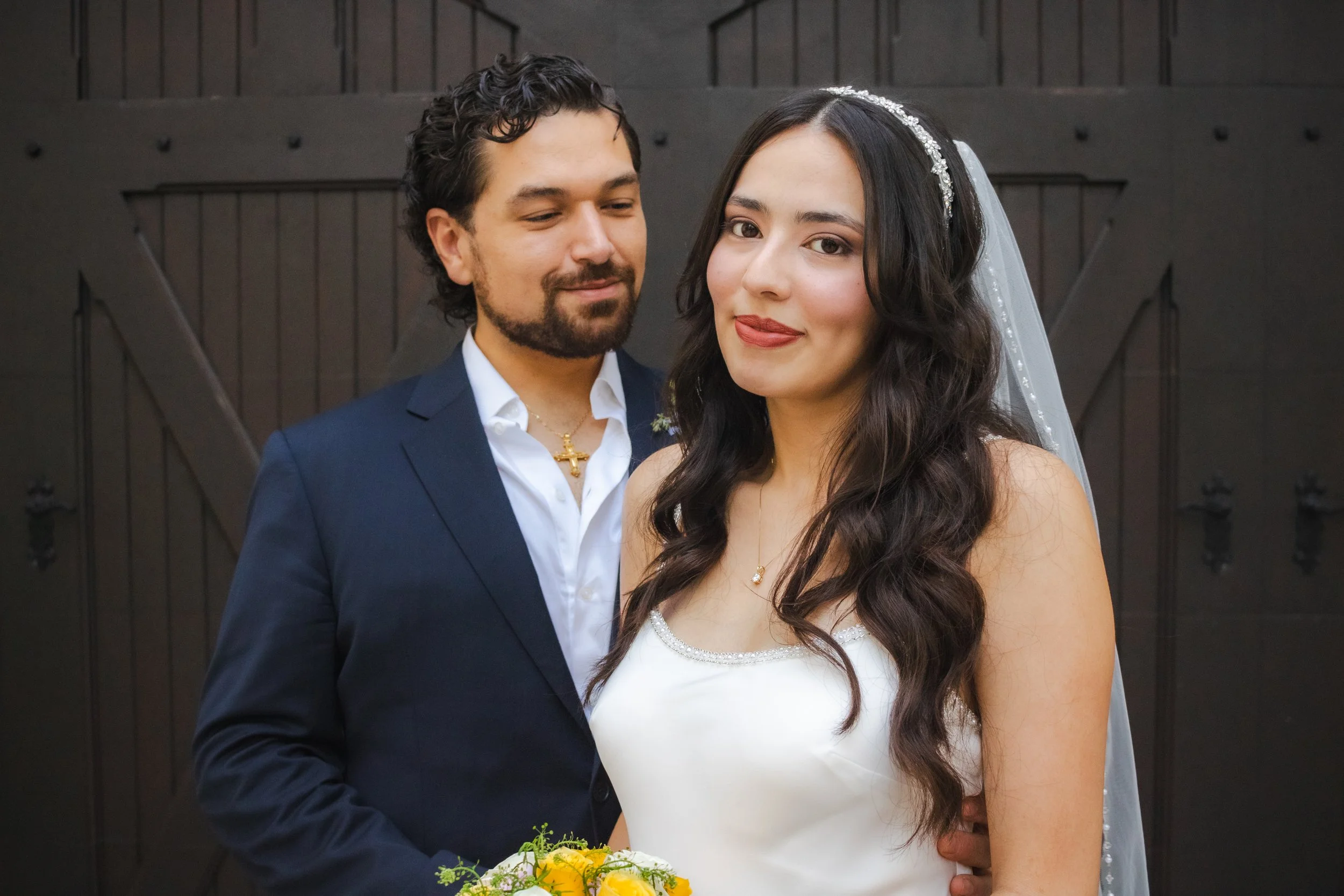 A bride and groom standing together on their wedding day in front of a dark wooden door. The groom is wearing a navy suit with a white shirt and a gold cross necklace, looking at her affectionately. The bride is wearing a white wedding dress with a b