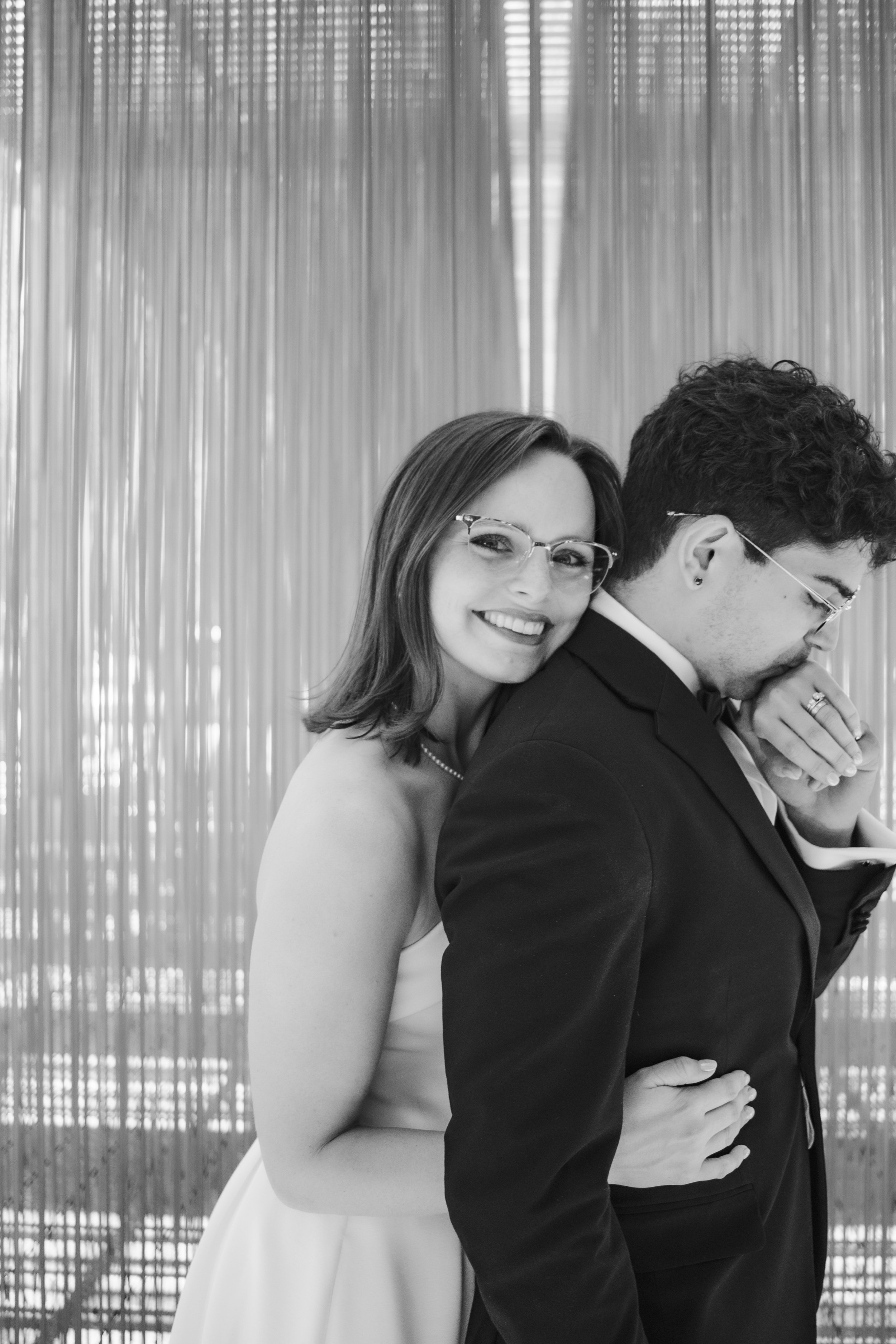 A smiling woman with glasses and a necklace, standing behind a man in a tuxedo with glasses, against a backdrop of vertical metallic bars.