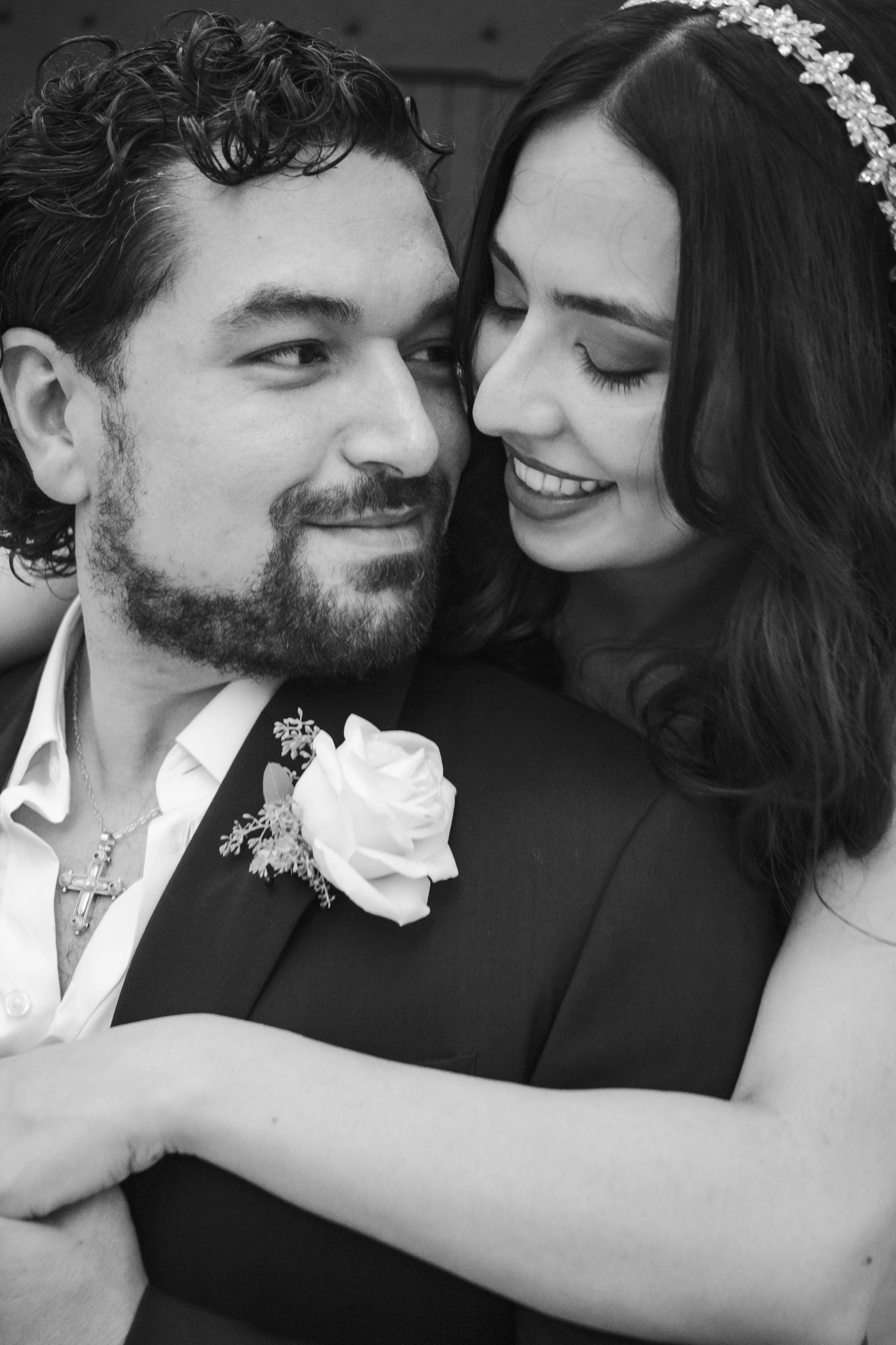 Close-up black and white photo of a couple at a wedding, smiling and looking into each other's eyes, with the woman's face partially hidden by her hair and head adorned with a floral headband.