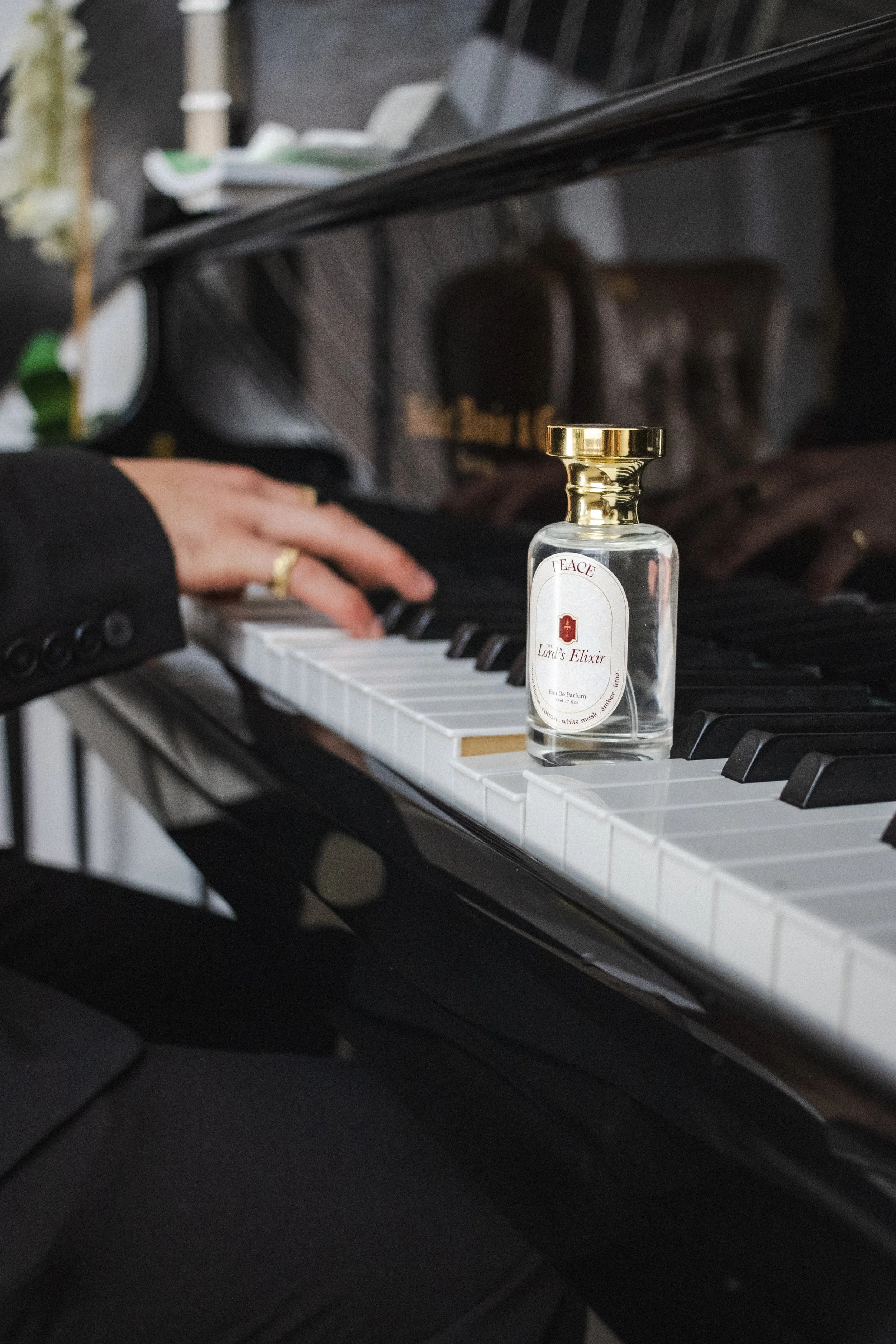 A person playing piano in a formal black suit with a ring on their finger, with a small glass bottle labeled "Lord's Elixir" placed on the piano keys.