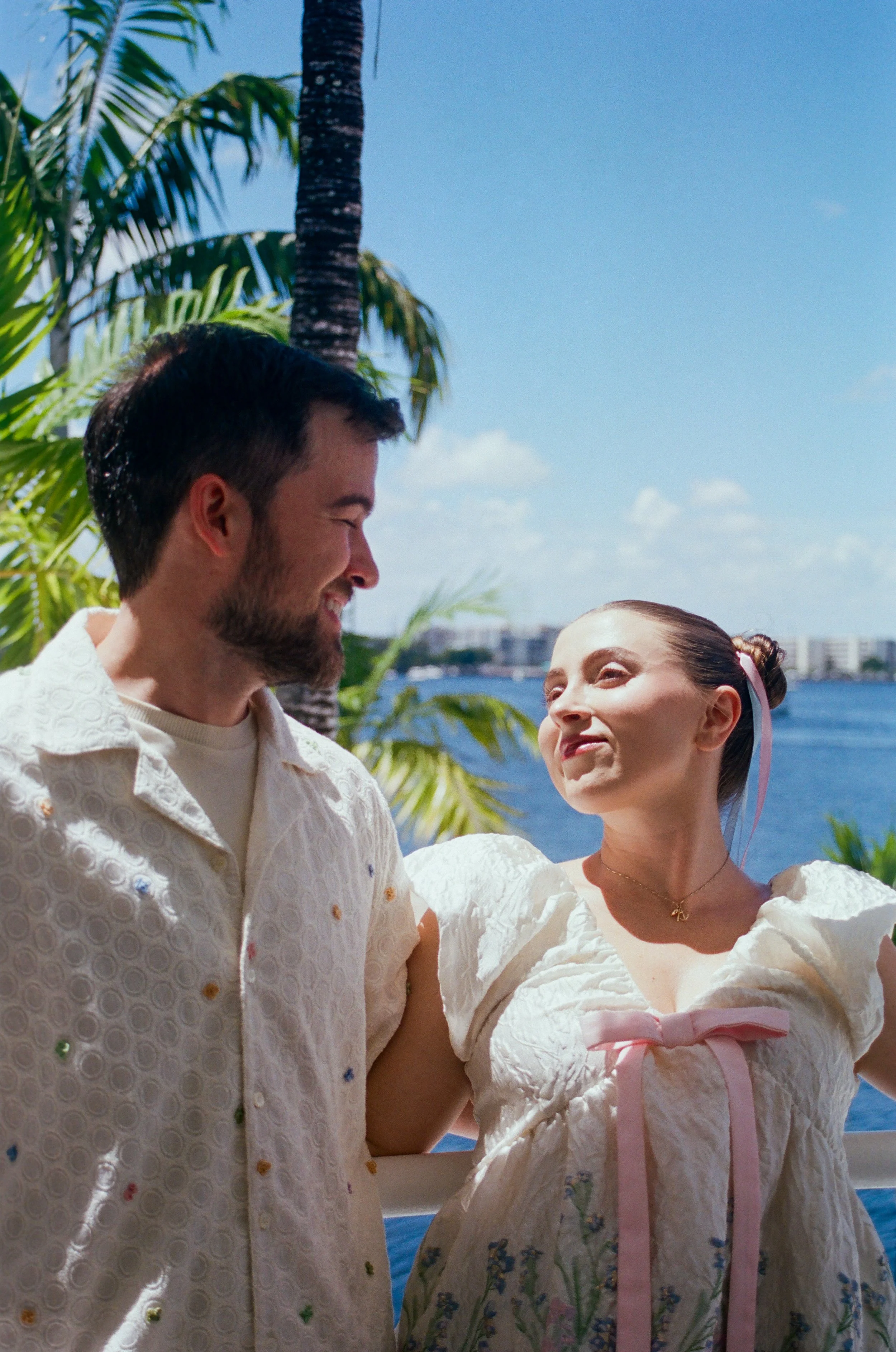 Two people, a man and a woman, are smiling and looking at each other outdoors near water, with palm trees and a city skyline in the background.