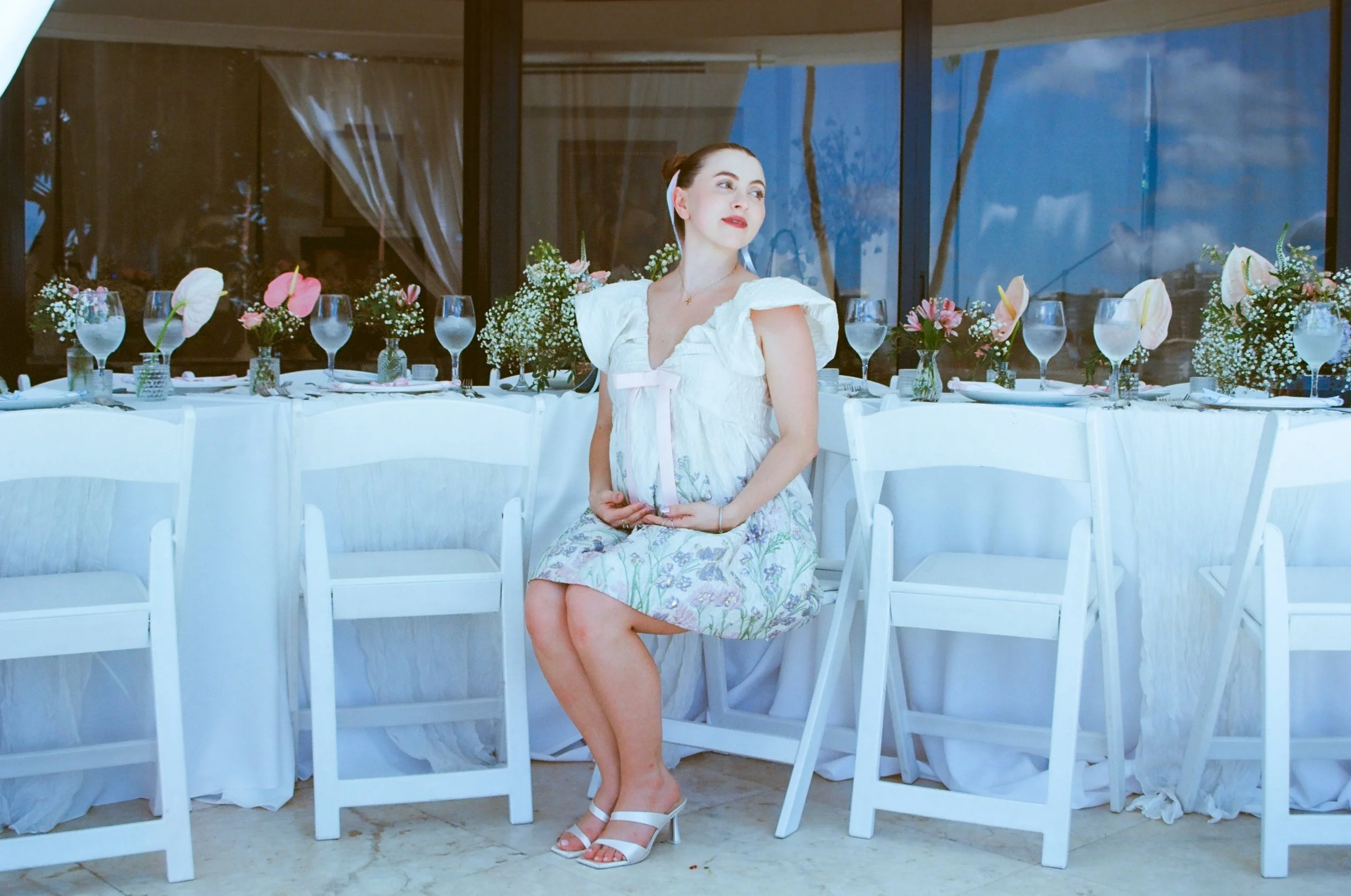 A young woman with dark hair in a bun wearing a white dress and white heels is sitting on a white chair at a decorated dining table with floral arrangements and glassware. She is looking to her right with a slight smile, in a room with large windows 